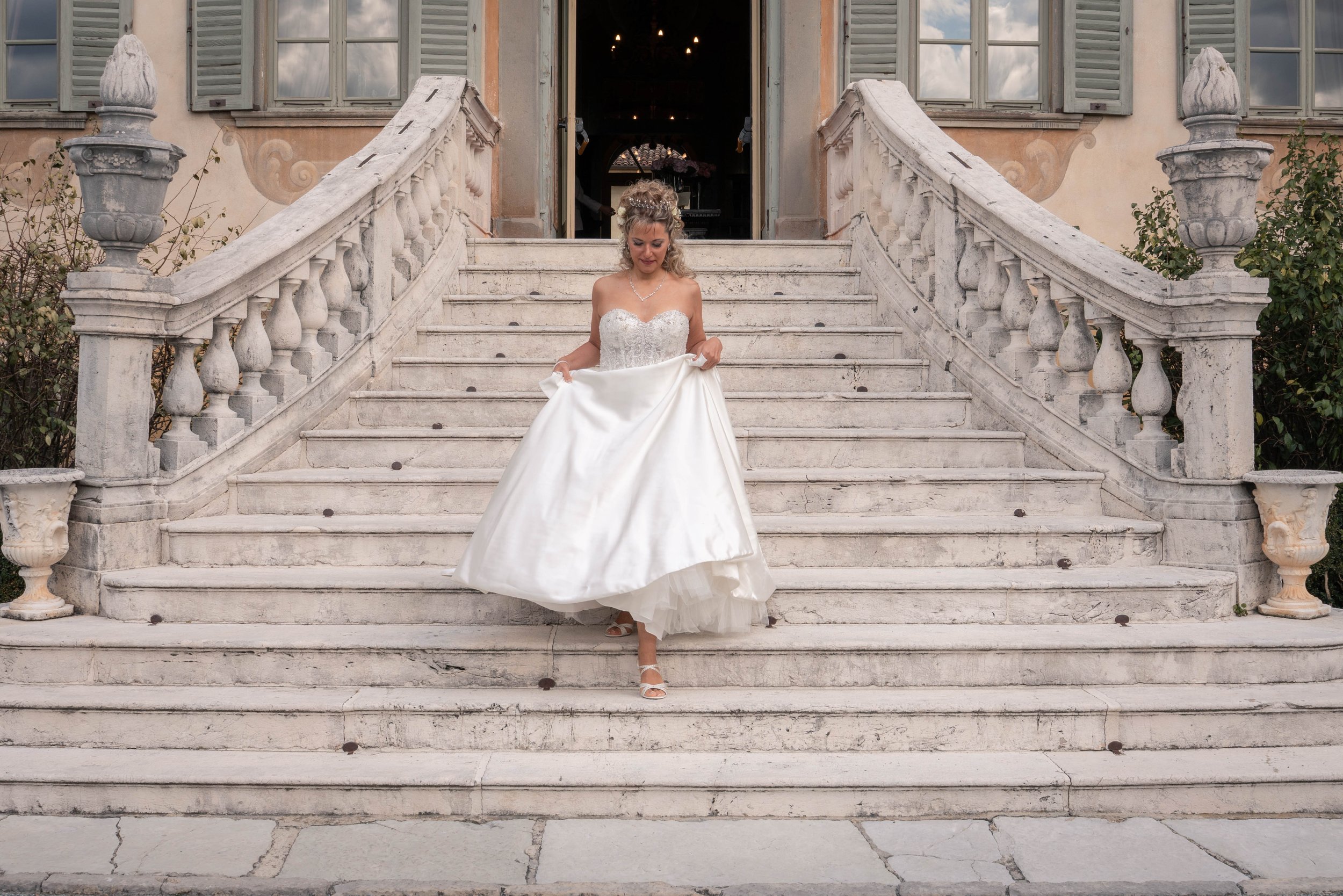 A bride in a white wedding dress standing on the stairs of a historic building with stone balustrades and green shutters. The bride is looking down and holding the skirt of her dress, with a necklace and floral headpiece.