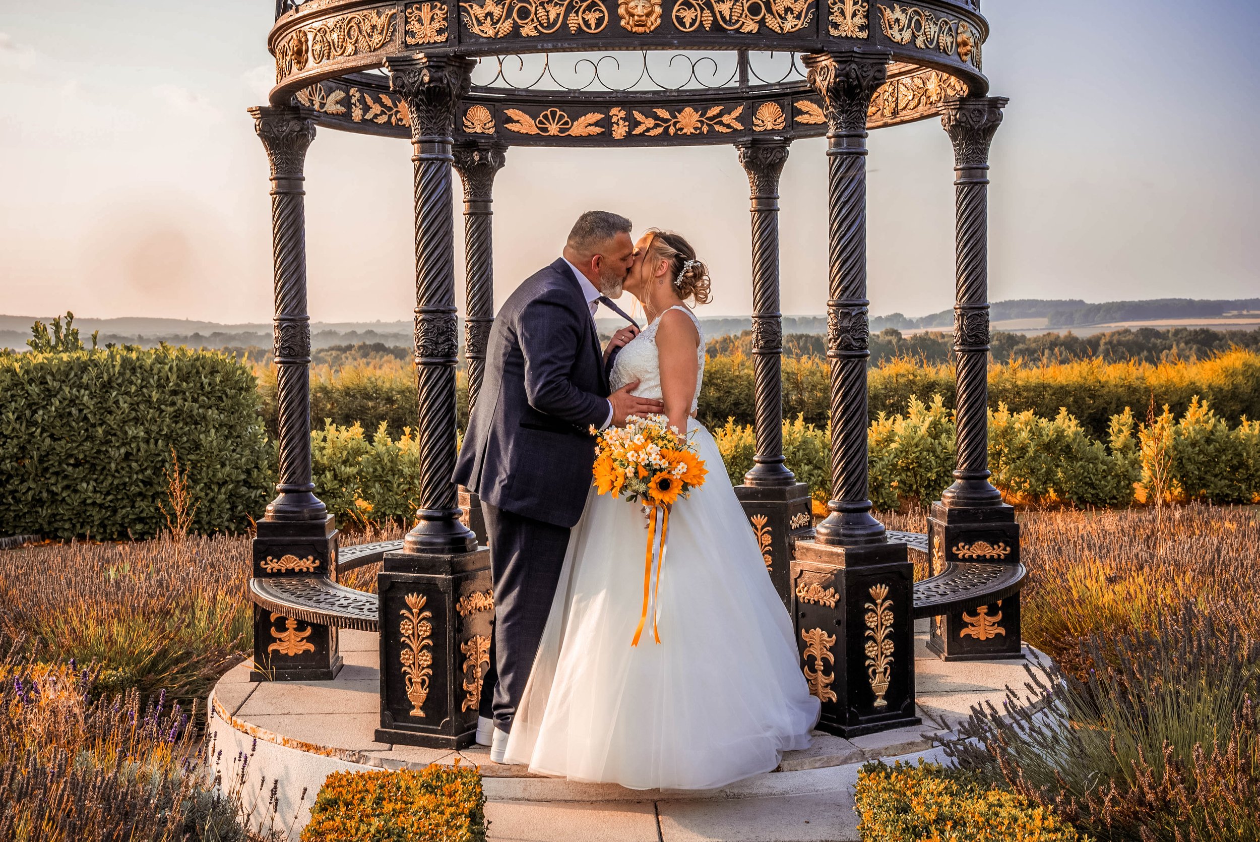 A bride and groom sharing a kiss under a decorative black and gold gazebo in an outdoor setting during sunset. The bride is holding a bouquet of sunflowers and other flowers, and the scenery includes fields and trees in the background.