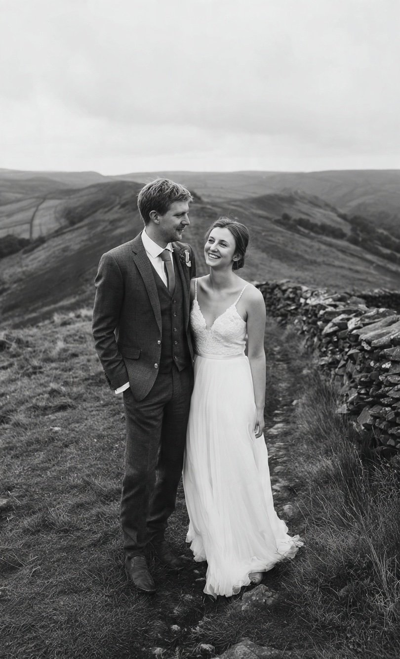 A smiling bride and groom walking together on a dirt path in a hilly landscape, with the bride in a white wedding dress and the groom in a suit.