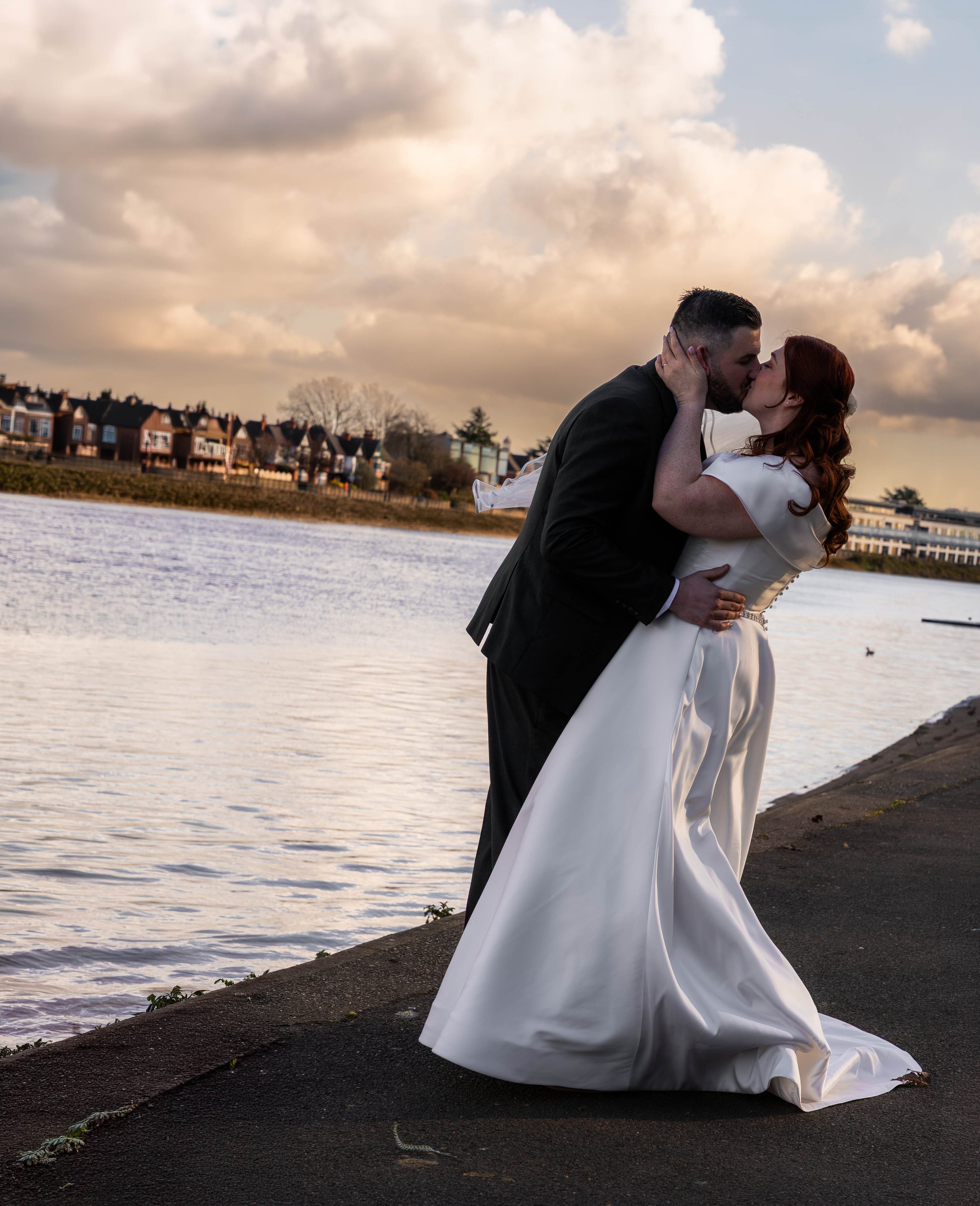 A bride and groom kissing along a riverbank at sunset.