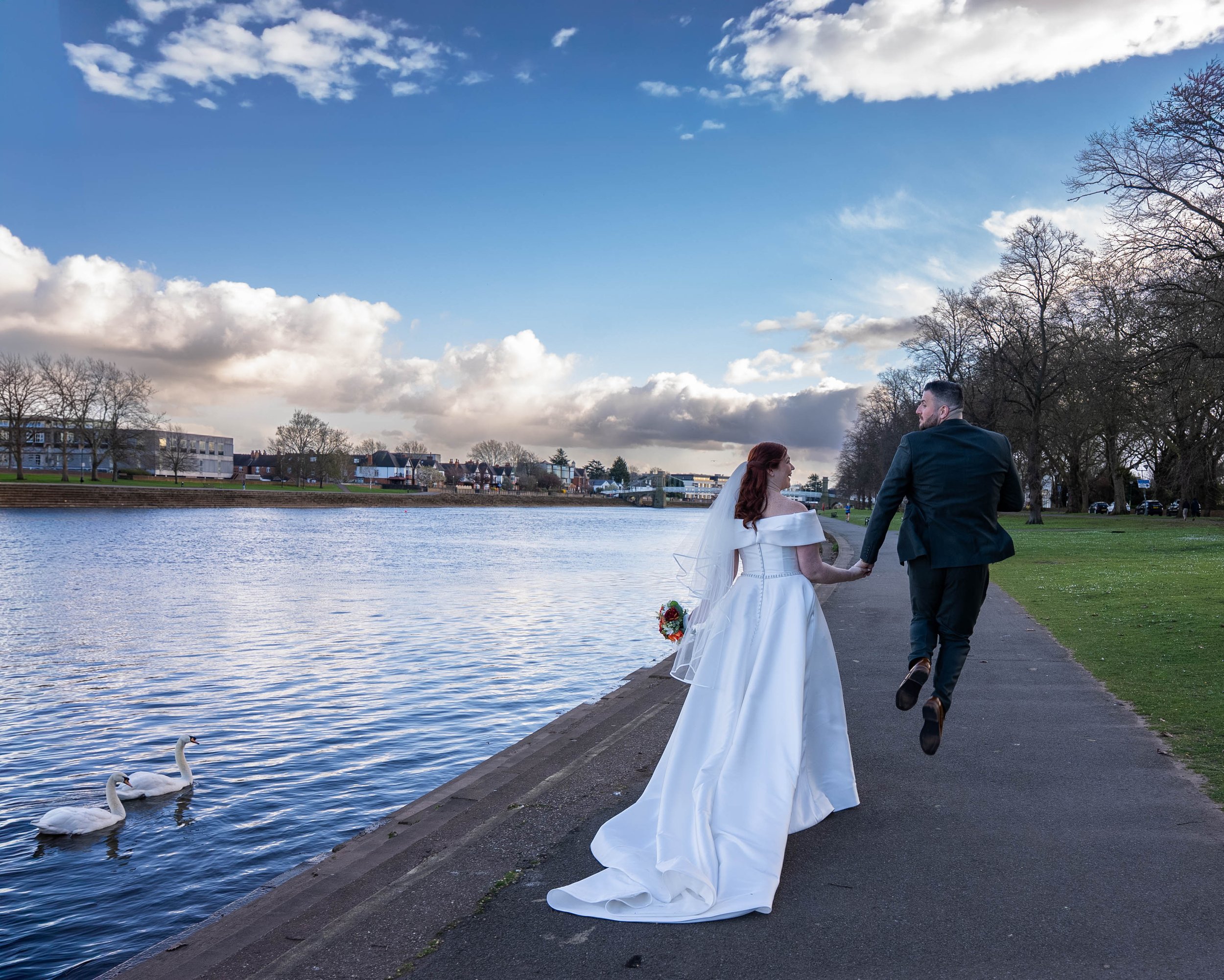 A bride and groom holding hands and running along a riverside pathway on their wedding day, with swans swimming in the river nearby and a cloudy sky above.