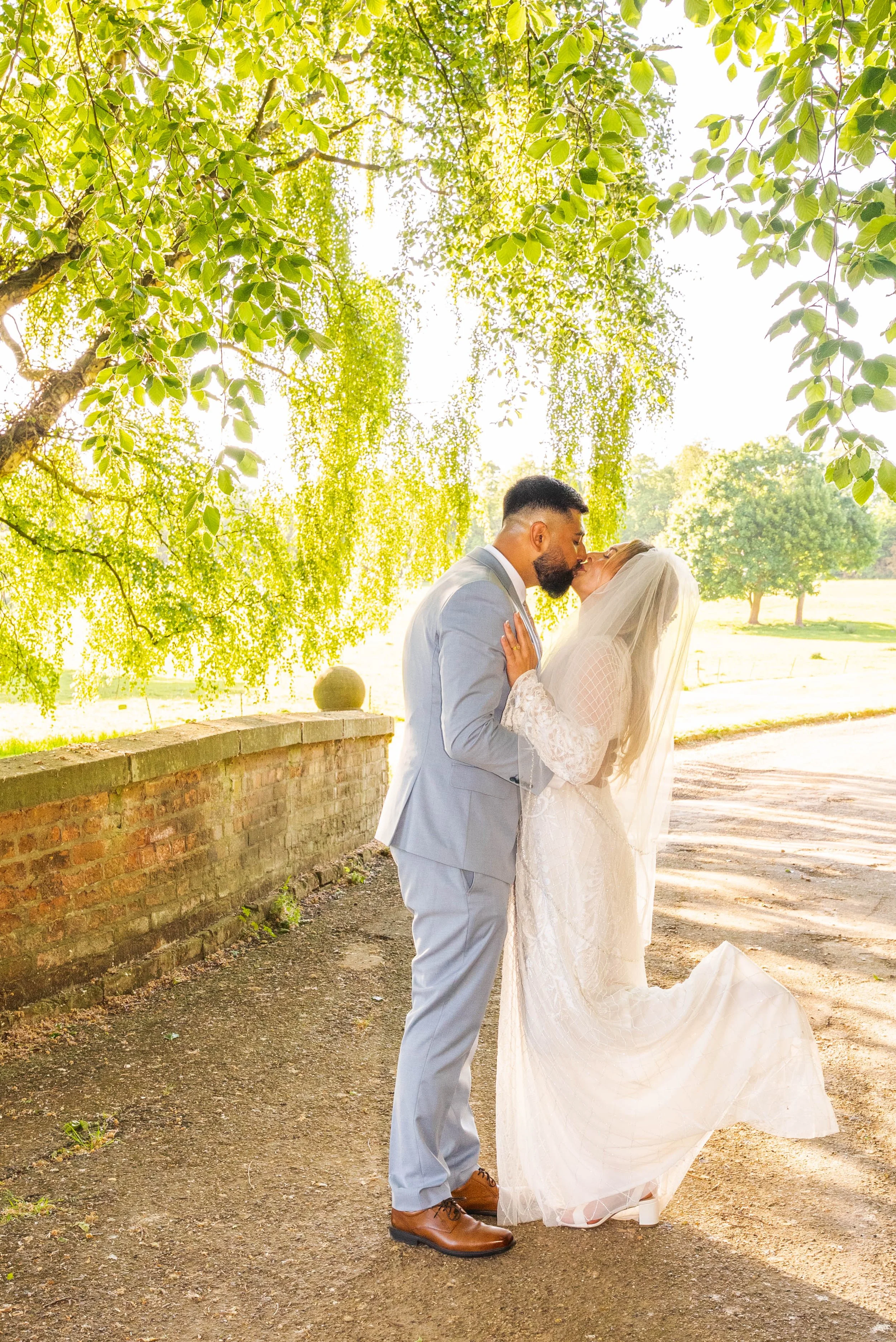 A newlywed couple sharing a kiss outdoors during sunset, surrounded by green trees and sunlight filtering through leaves.
