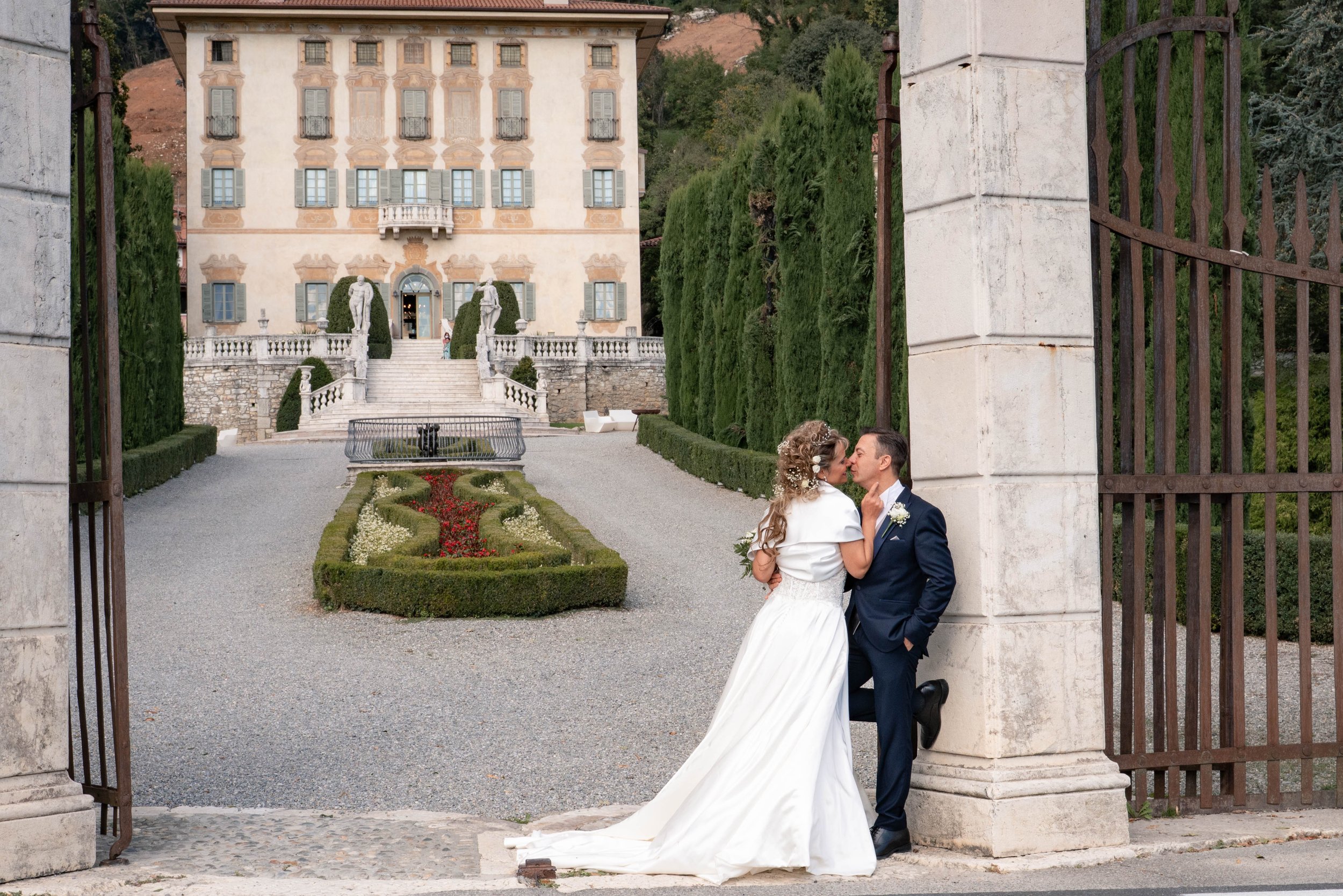 A bride and groom standing close together, smiling, in front of a large ornate house with a staircase and garden, viewed through an open wrought iron gate.