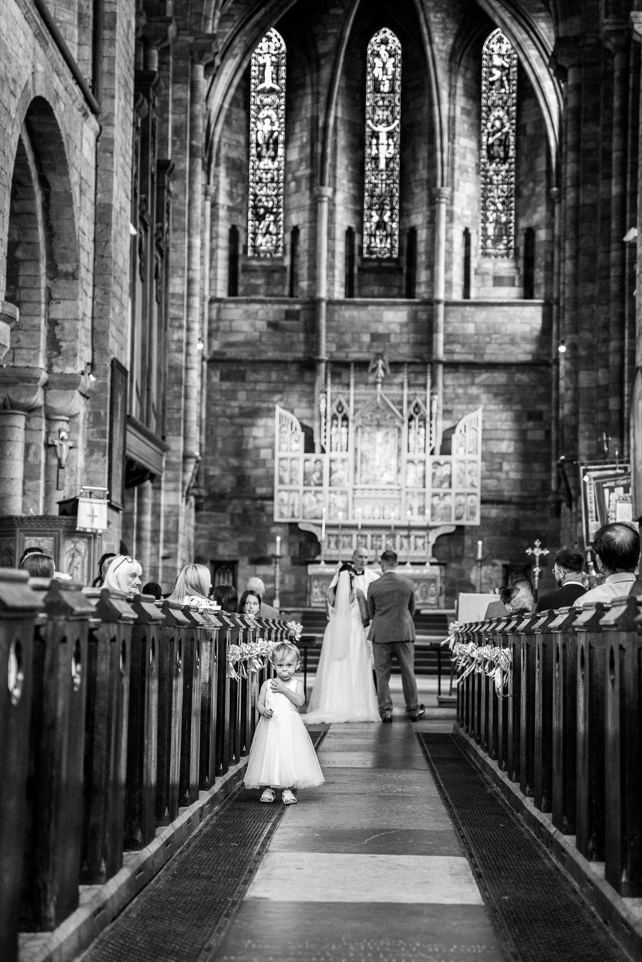 A wedding ceremony taking place in a church with a young flower girl standing in the aisle, a bride and groom at the altar, stained glass windows, and pews decorated with ribbons.