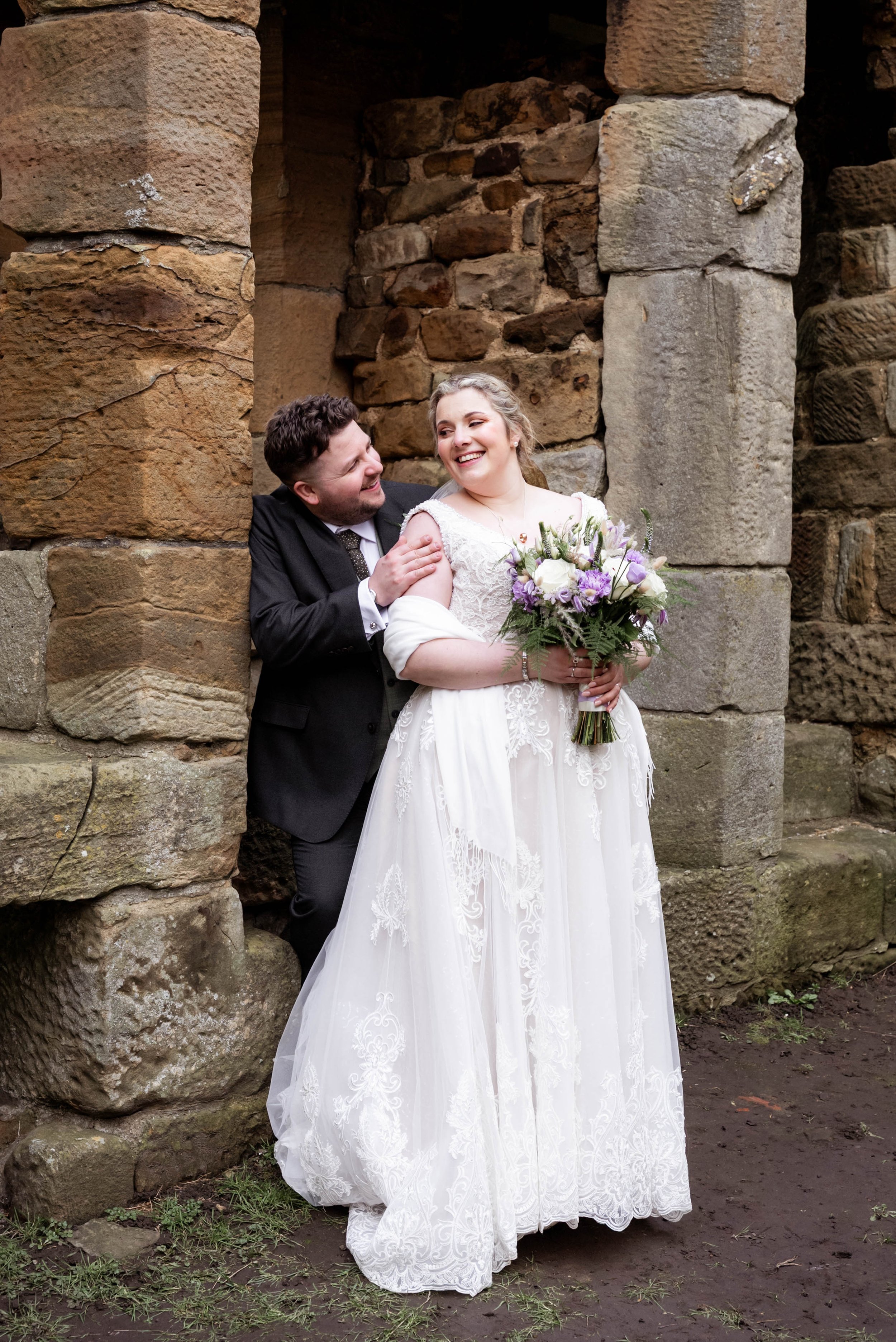 A bride and groom laughing and smiling together during their wedding, standing outdoors against a stone wall. The bride is holding a bouquet of flowers and wearing a white lace wedding dress, while the groom is dressed in a black suit with a vest and
