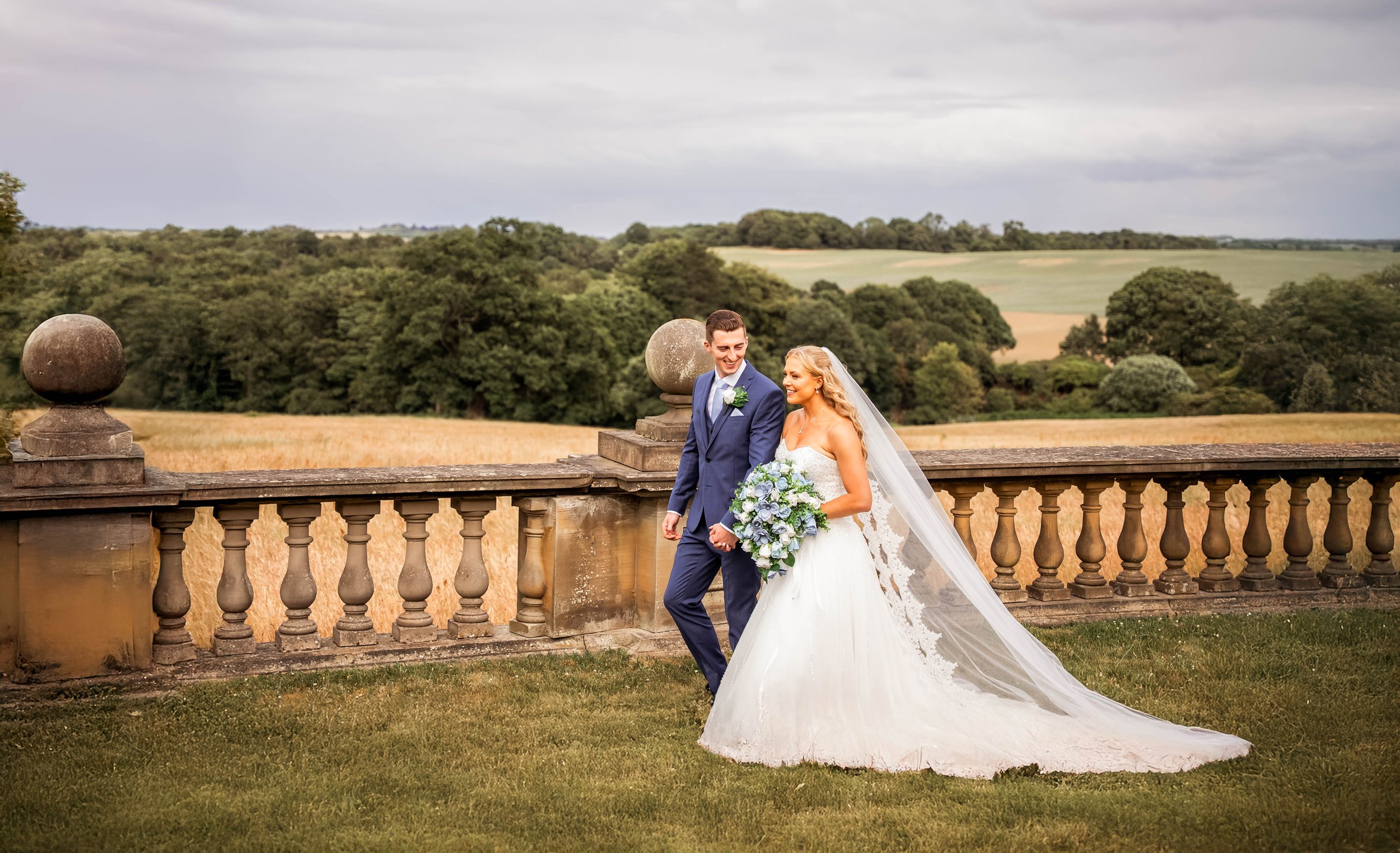 A newlywed couple holding hands and smiling, standing outdoors on a stone balcony overlooking a lush green landscape with fields and trees under a cloudy sky.