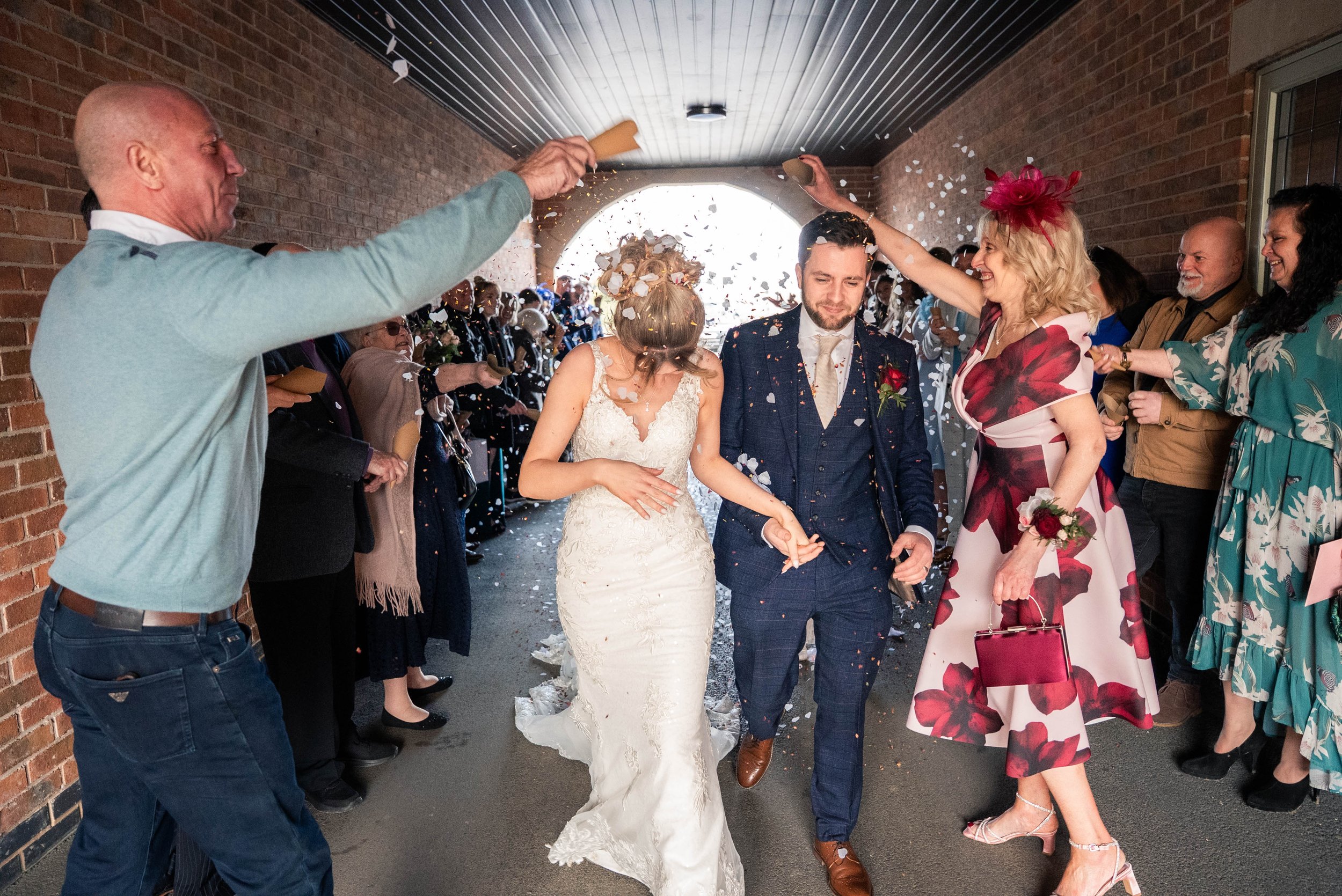 Bride and groom walking through a hallway lined with guests throwing confetti to celebrate. The bride is wearing a white lace wedding dress and the groom is in a blue checkered suit. Guests are smiling and throwing confetti, with some women dressed i