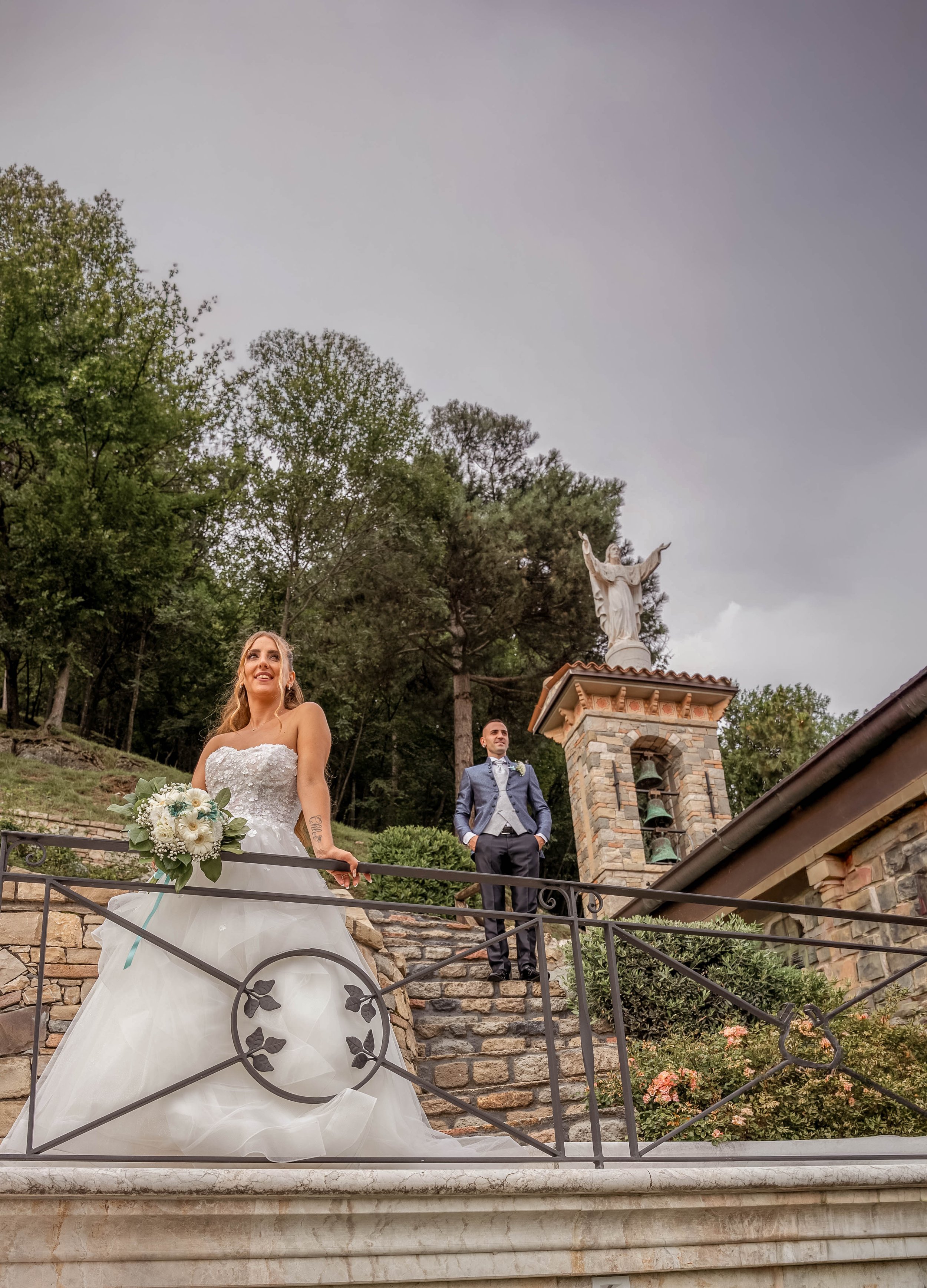 A bride holding a bouquet of white flowers and a groom standing on a stone staircase outdoors near a religious statue of Jesus Christ in front of trees, under a cloudy sky.