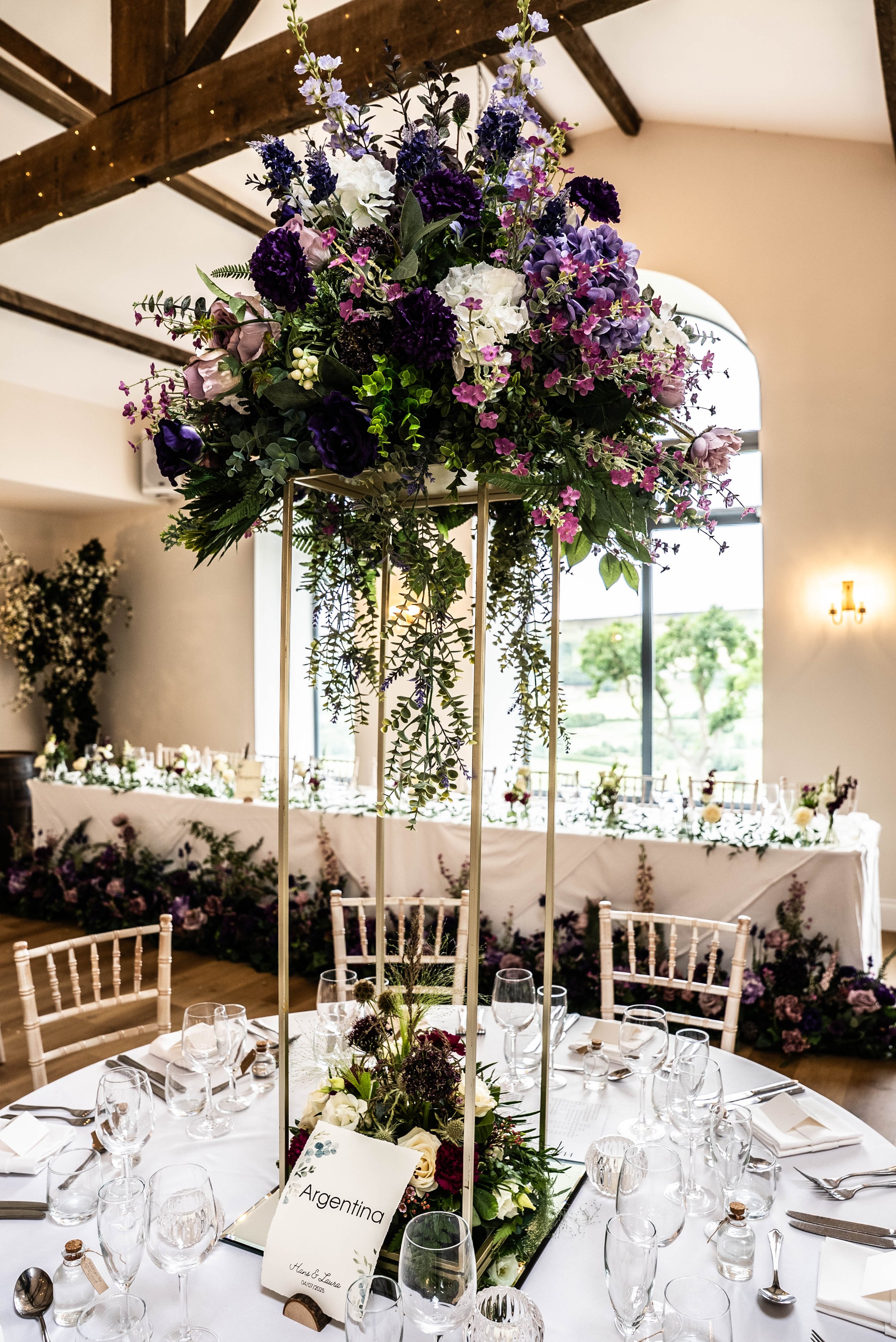 A tall centerpiece floral arrangement with purple, white, and pink flowers on a dining table at an event venue, with a large window in the background and a long decorated table nearby.