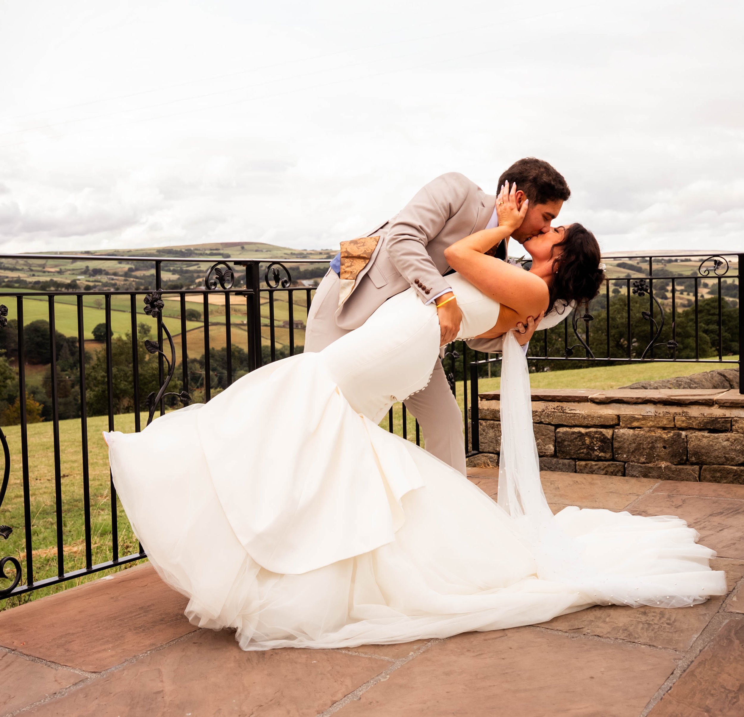 A bride and groom sharing a kiss on a balcony, with the groom dipping the bride. The bride is in a white wedding gown and the groom in a light-colored suit against a rural landscape background.