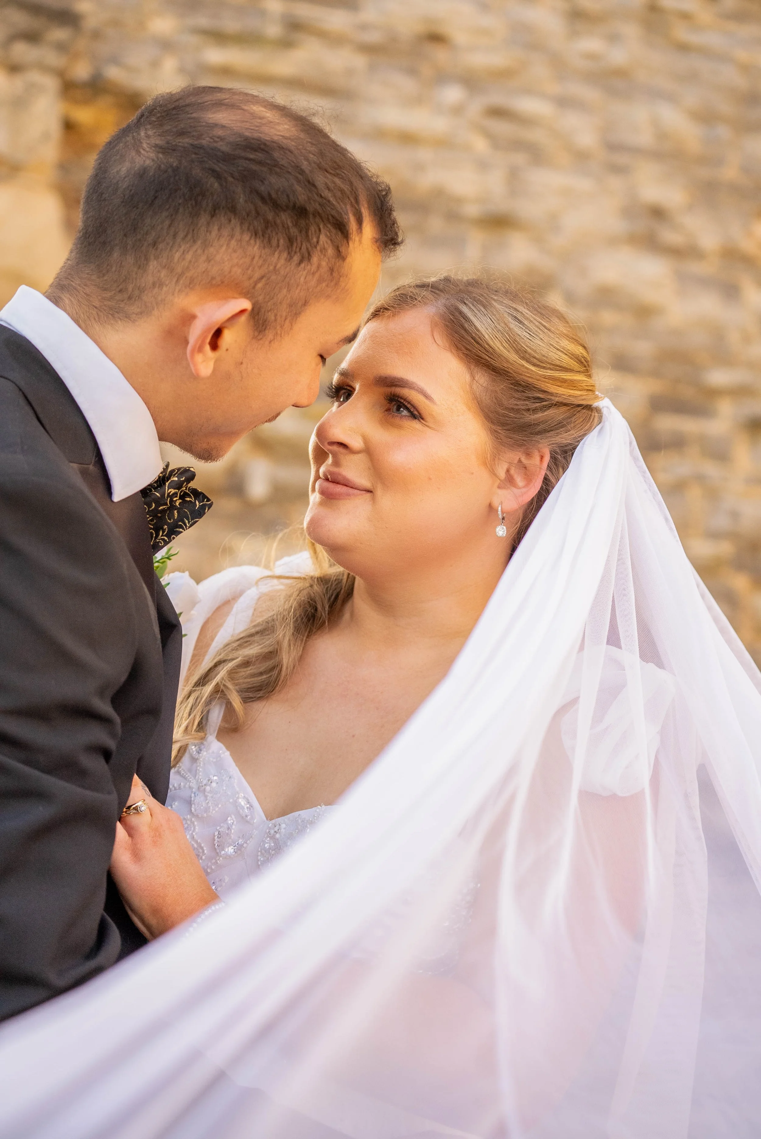 A bride and groom gazing into each other's eyes during their wedding, with a rustic stone wall in the background.