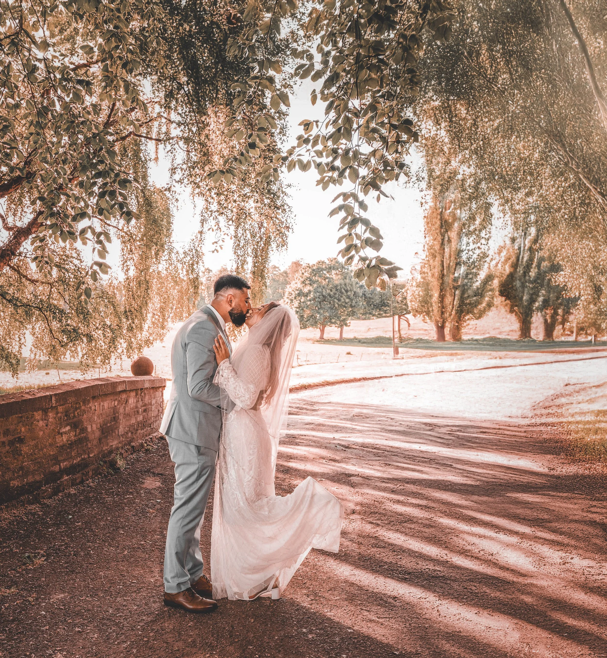A bride and groom sharing a kiss outdoors under a tree, with sunlight filtering through the leaves. The bride is wearing a white wedding dress and veil, and the groom is dressed in a light gray suit.