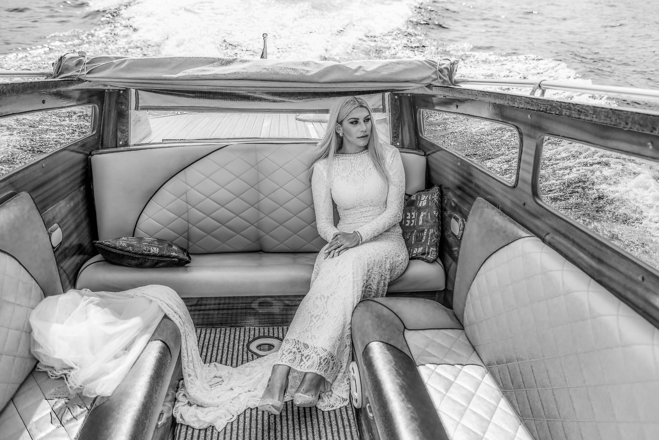 A woman with long hair in a lace dress sitting on a leather and wooden bench inside a boat, with water visible outside the boat windows.