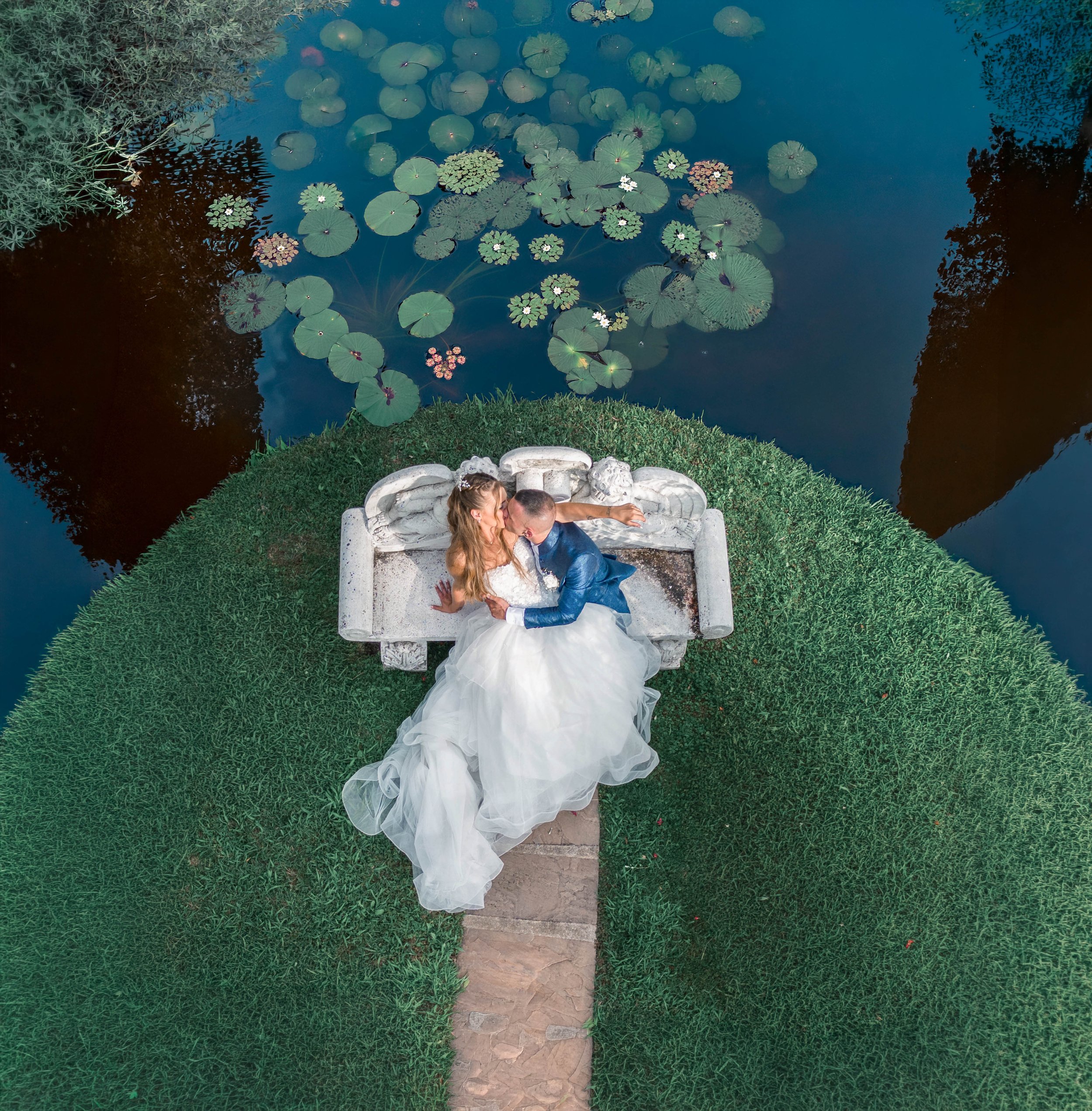 A bride and groom sitting on a stone bench by a pond with lily pads and white flowers, surrounded by greenery.