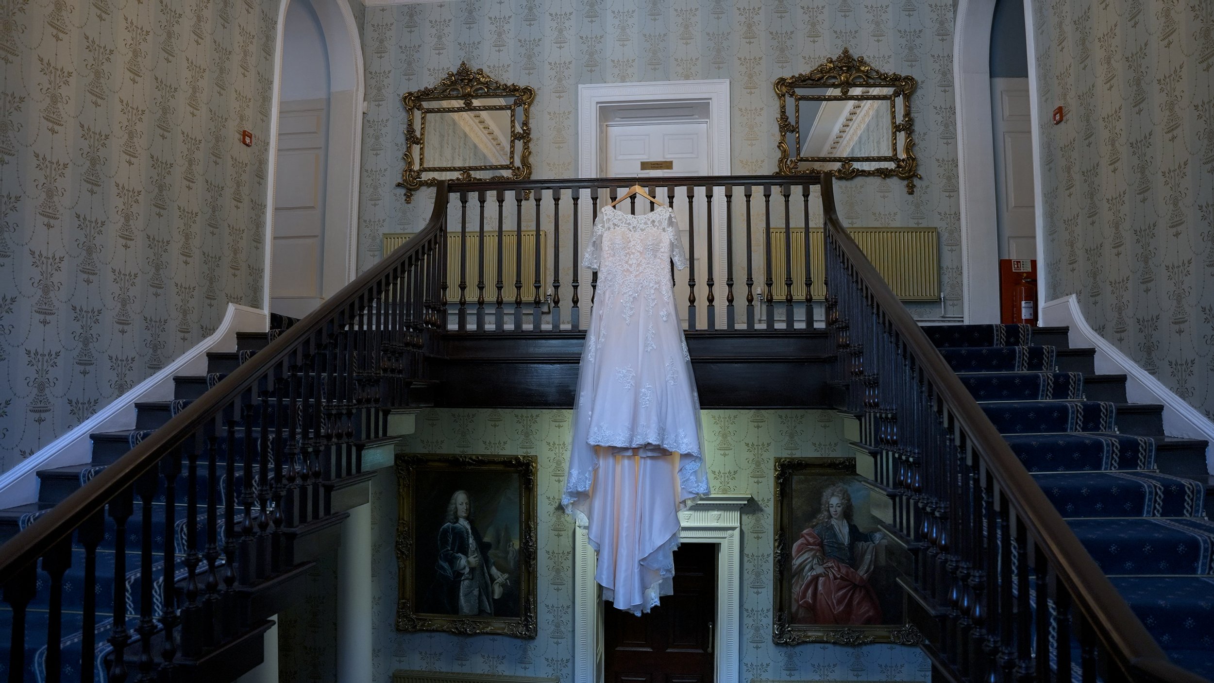White wedding dress hanging on a hanger centered on a staircase railing in an elegant, vintage-style interior with patterned wallpaper, ornate gold-framed mirrors, and classic portrait paintings.