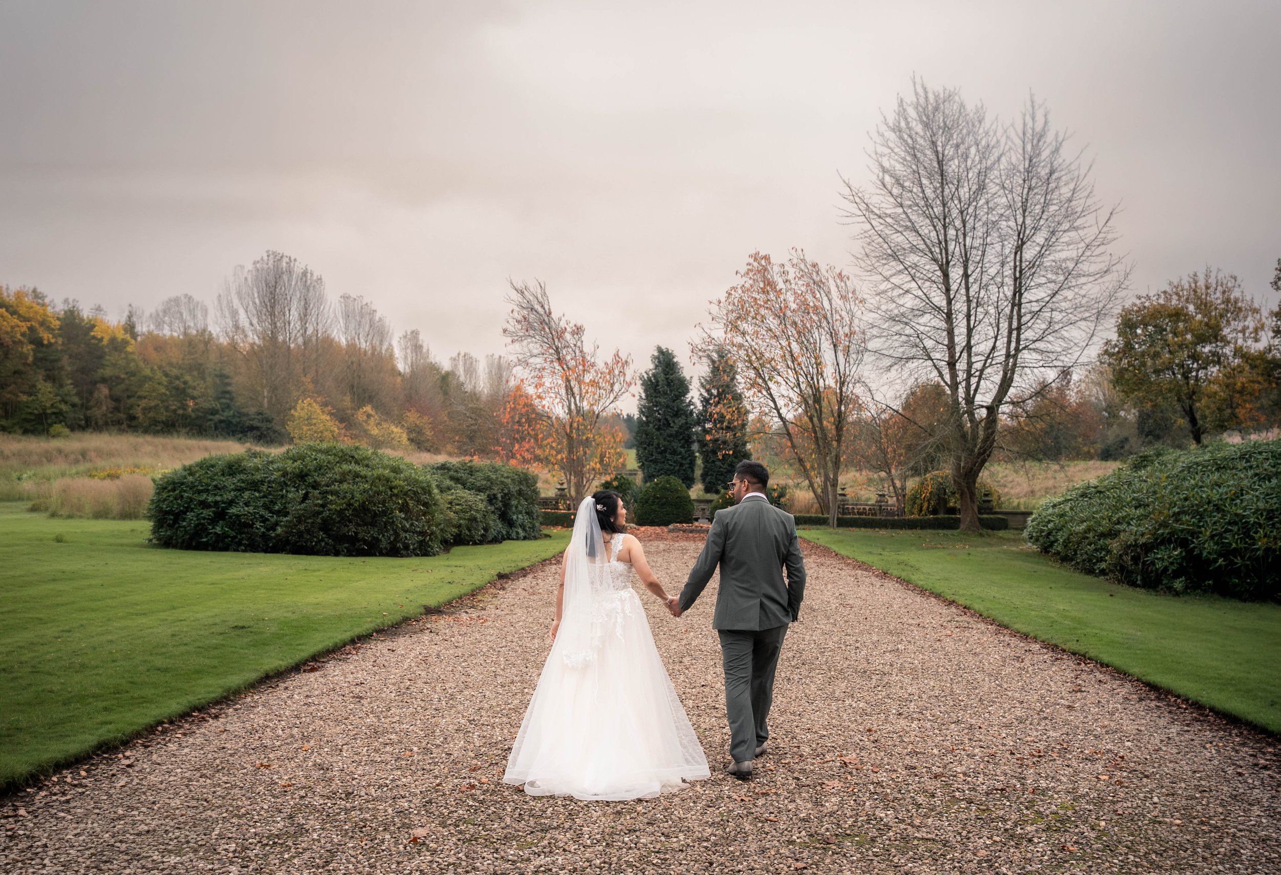 A bride and groom holding hands and walking on a gravel path through a park with trees and bushes, during autumn with overcast sky.