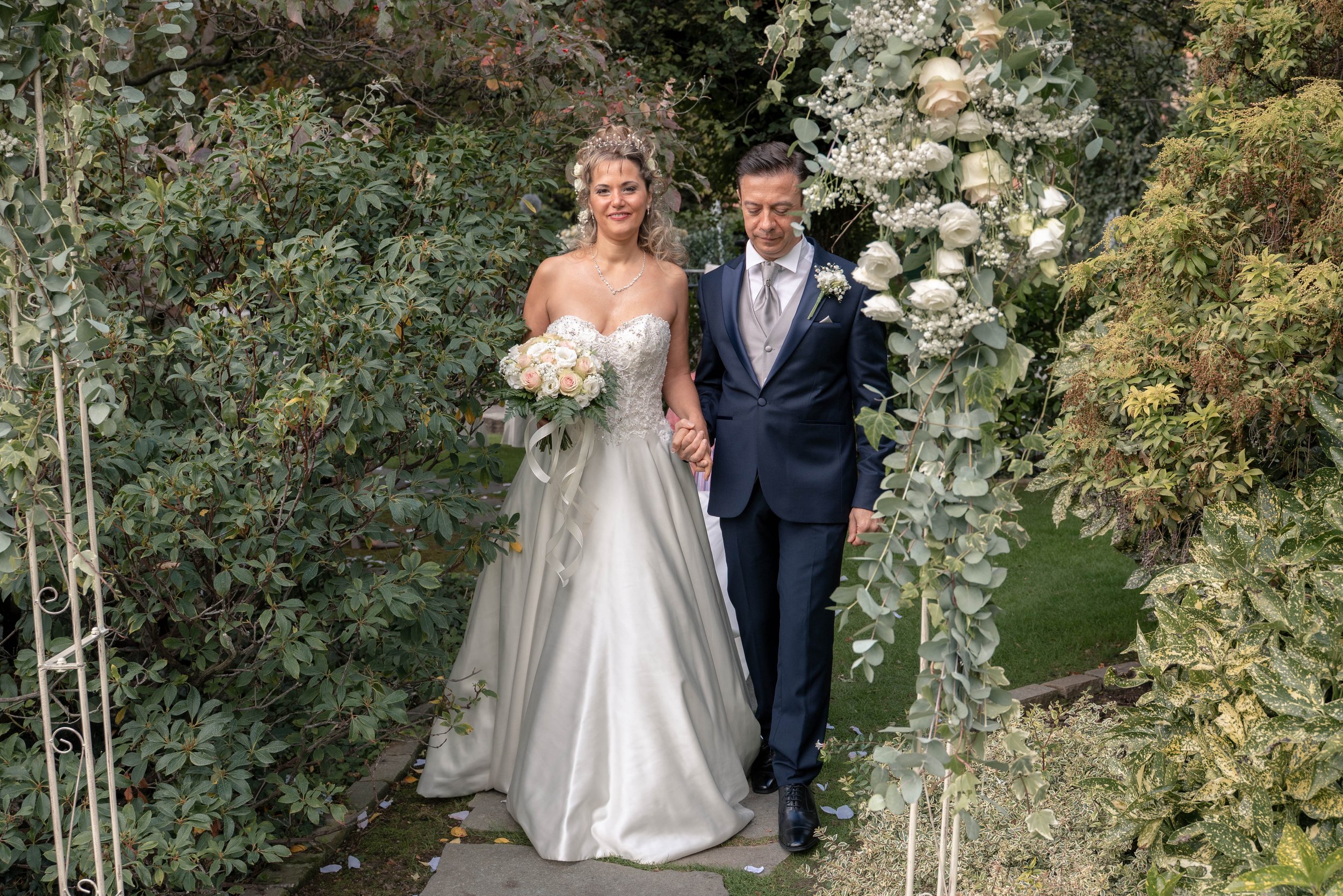 A bride and groom holding hands walking through an archway decorated with white roses, greenery, and baby's breath during a wedding ceremony outdoors.