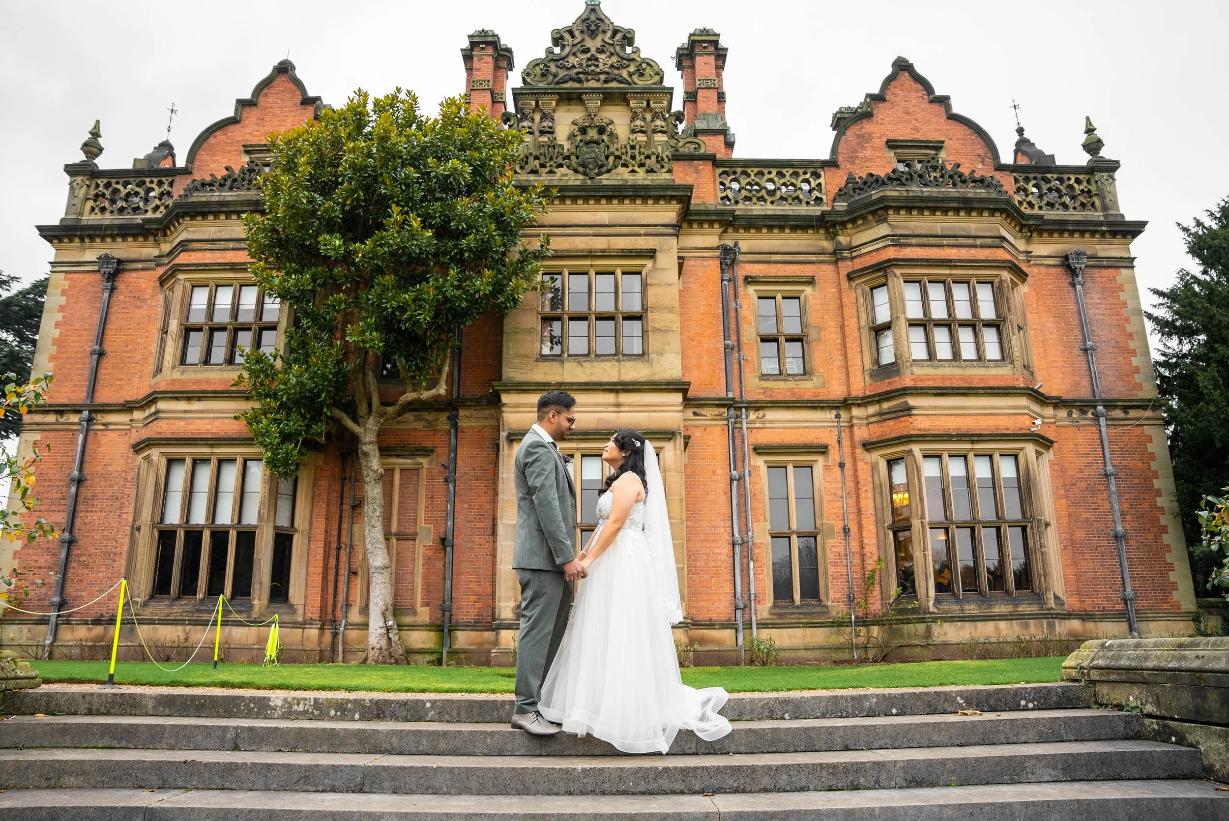 A bride and groom holding hands and gazing at each other in front of a historic brick building with large windows and ornate architectural details, on a set of stone stairs.