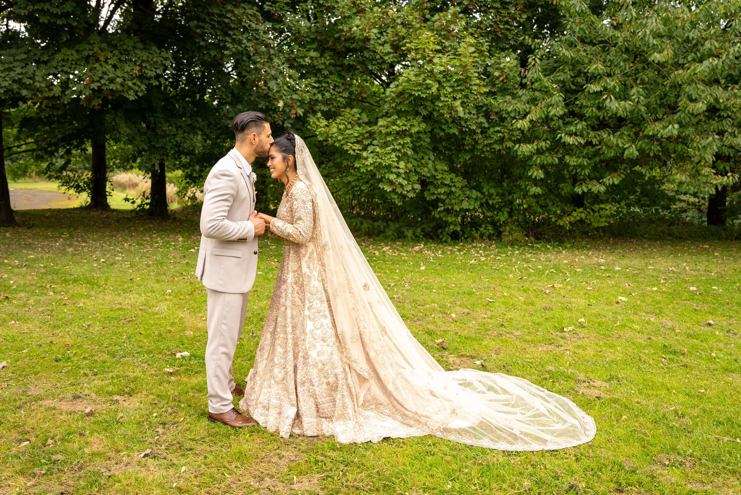 A bride and groom holding hands and smiling at each other outdoors in a park with green trees in the background.