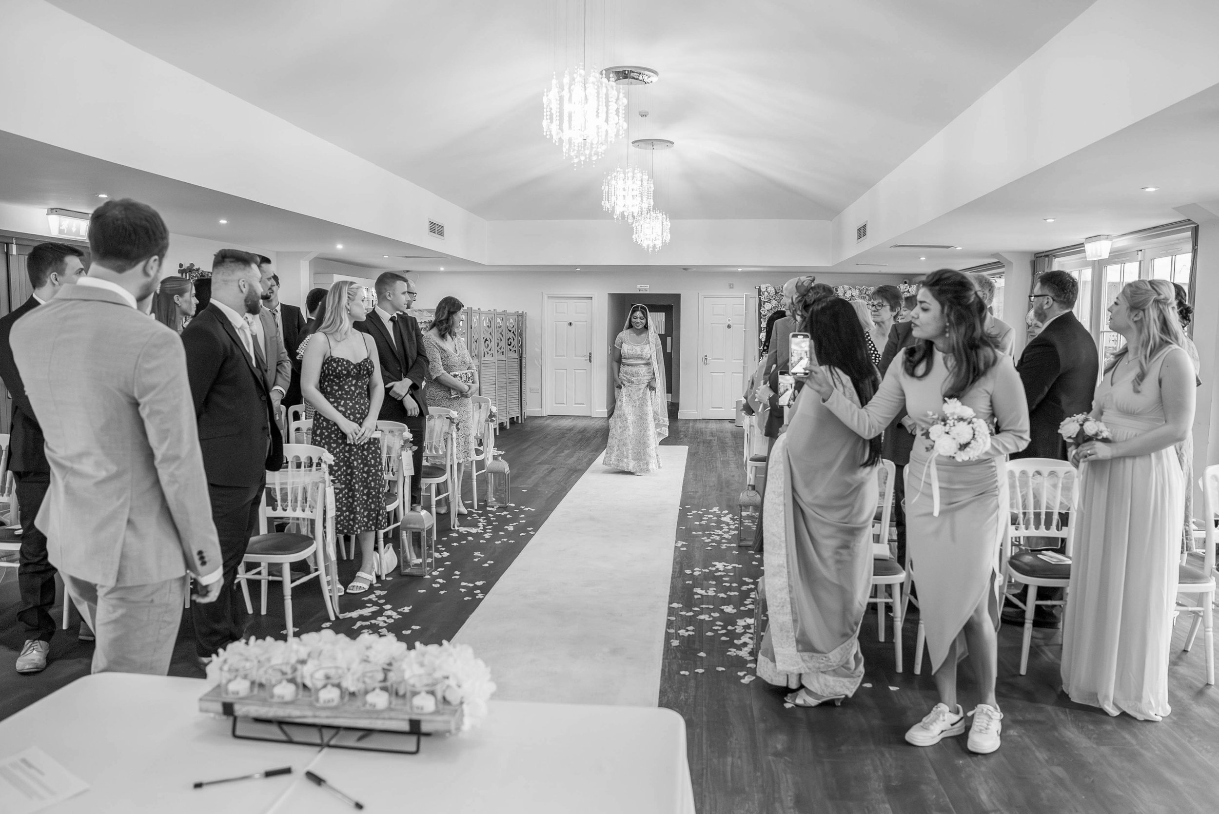A black-and-white photo of a wedding ceremony with the bride walking down the aisle in a bridal gown and veil. Guests are standing on both sides, some taking photos, in a decorated indoor venue with chandeliers and flower petal scattered on the floor