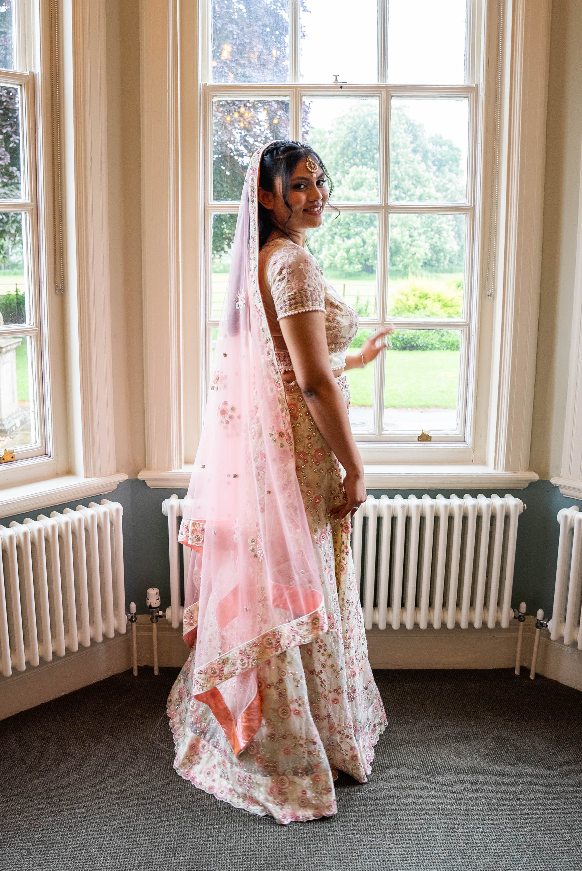 A woman dressed in a traditional South Asian bridal outfit standing by a window.