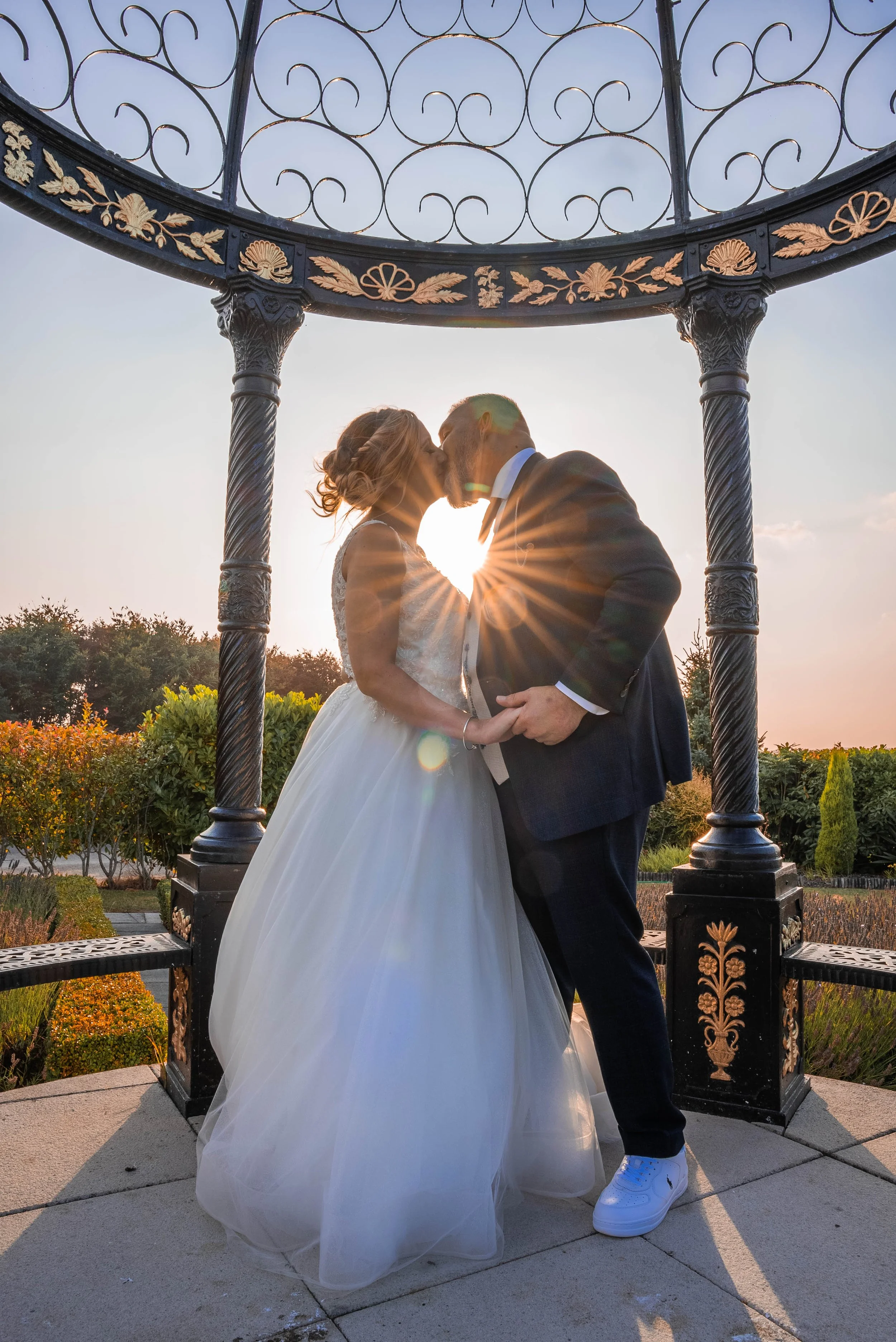A bride and groom sharing a kiss during sunset under an ornate black and gold gazebo, with the sun creating a starburst effect between them.