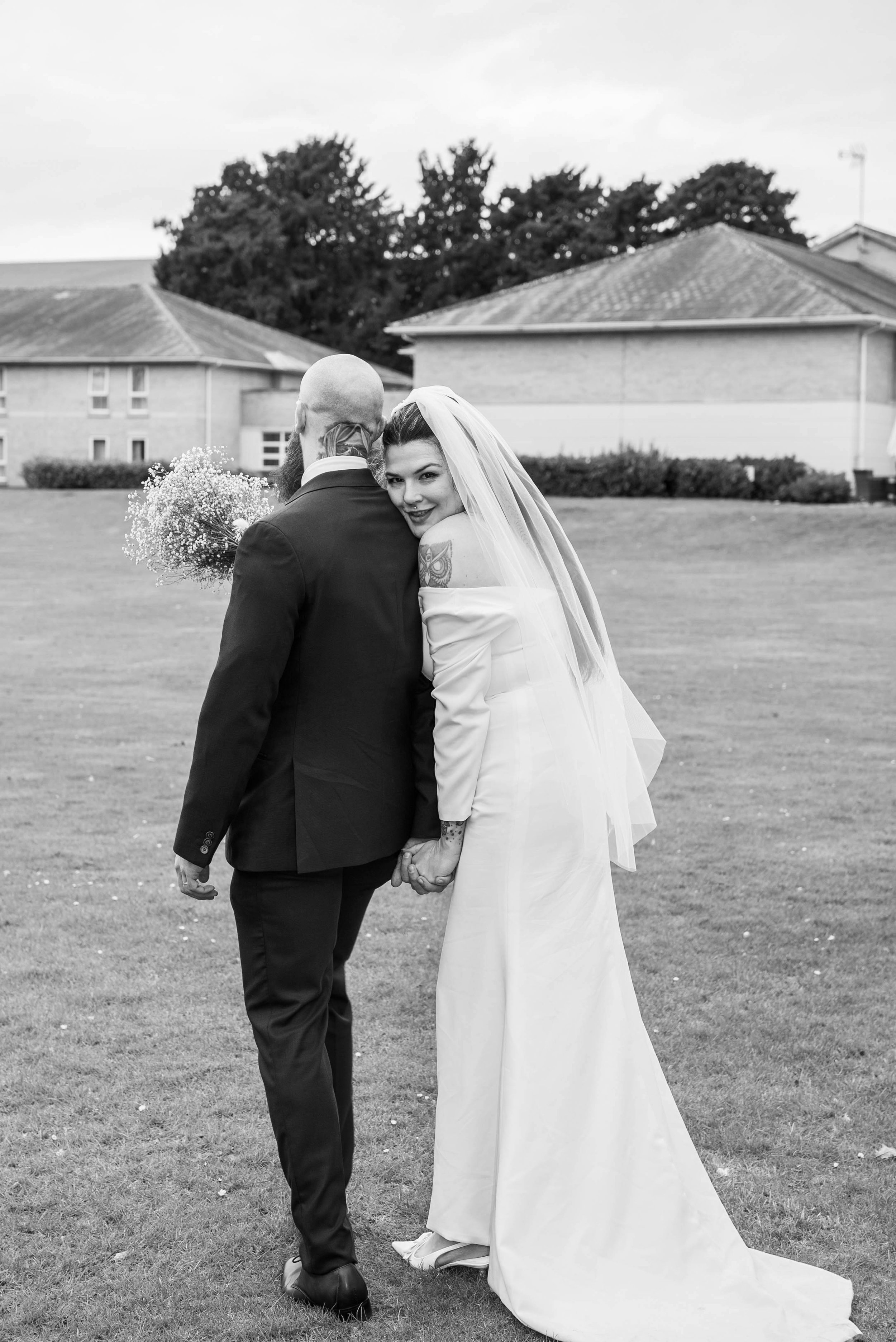 A bride and groom holding hands outdoors on a grassy area, facing each other and smiling, with trees and residential buildings in the background.