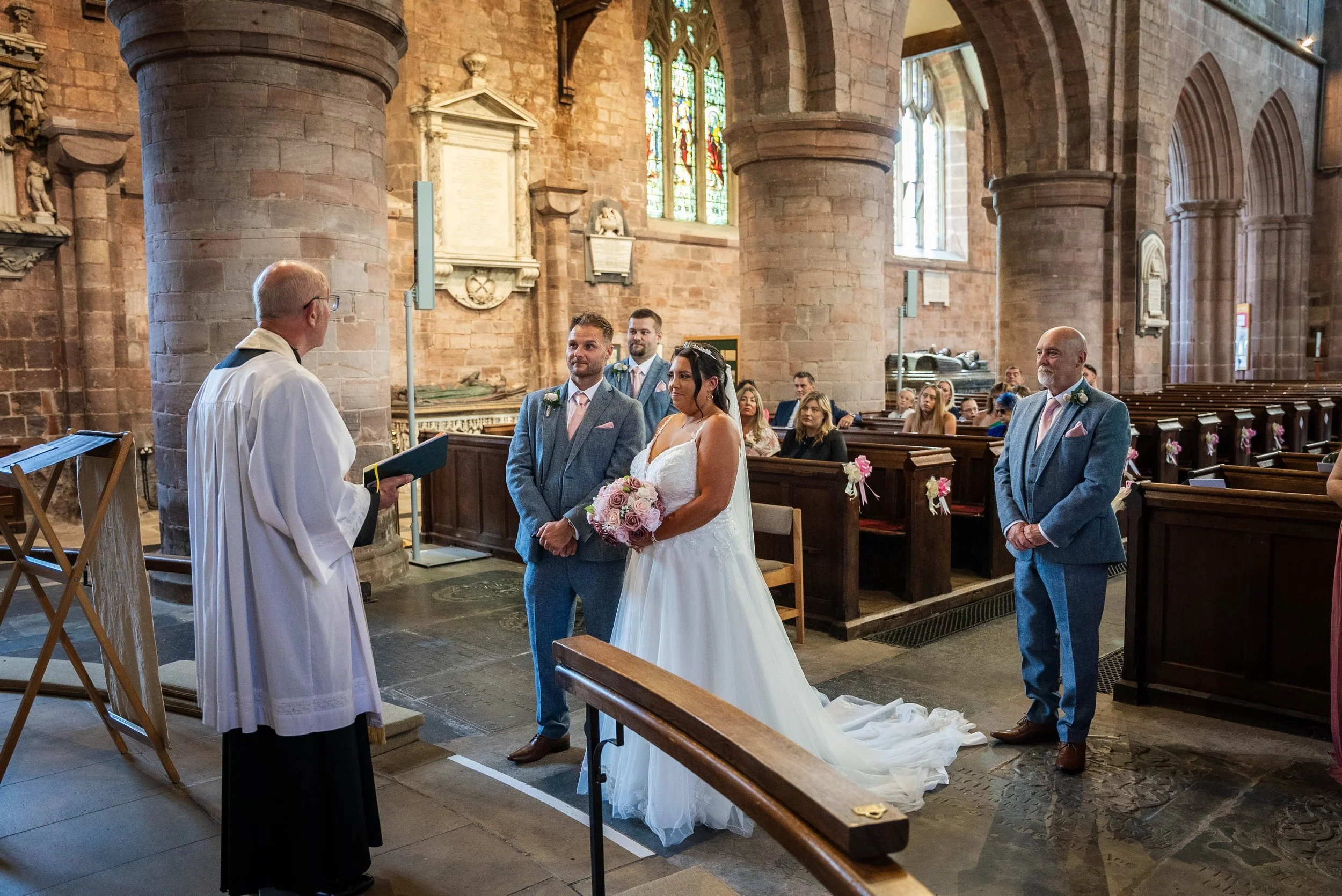 A wedding ceremony taking place inside a stone church with stained glass windows, where a priest is officiating in front of a bride and groom, with the bride holding a bouquet and the groom standing beside her, surrounded by family and friends.