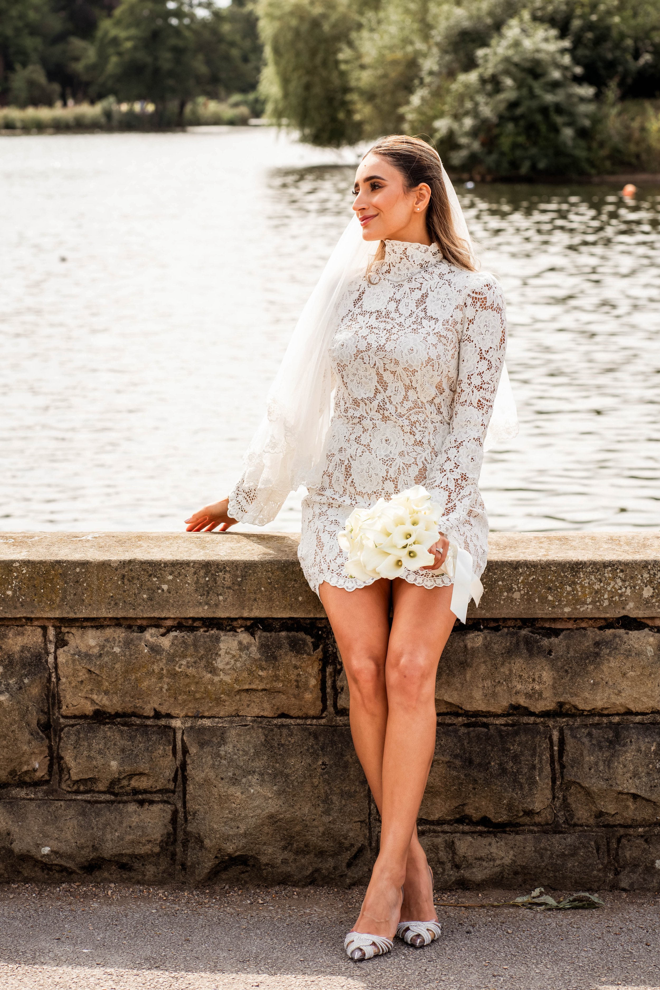 Woman dressed in a lace bridal gown holding a bouquet of white calla lilies, standing by a stone ledge next to a river with trees in the background.