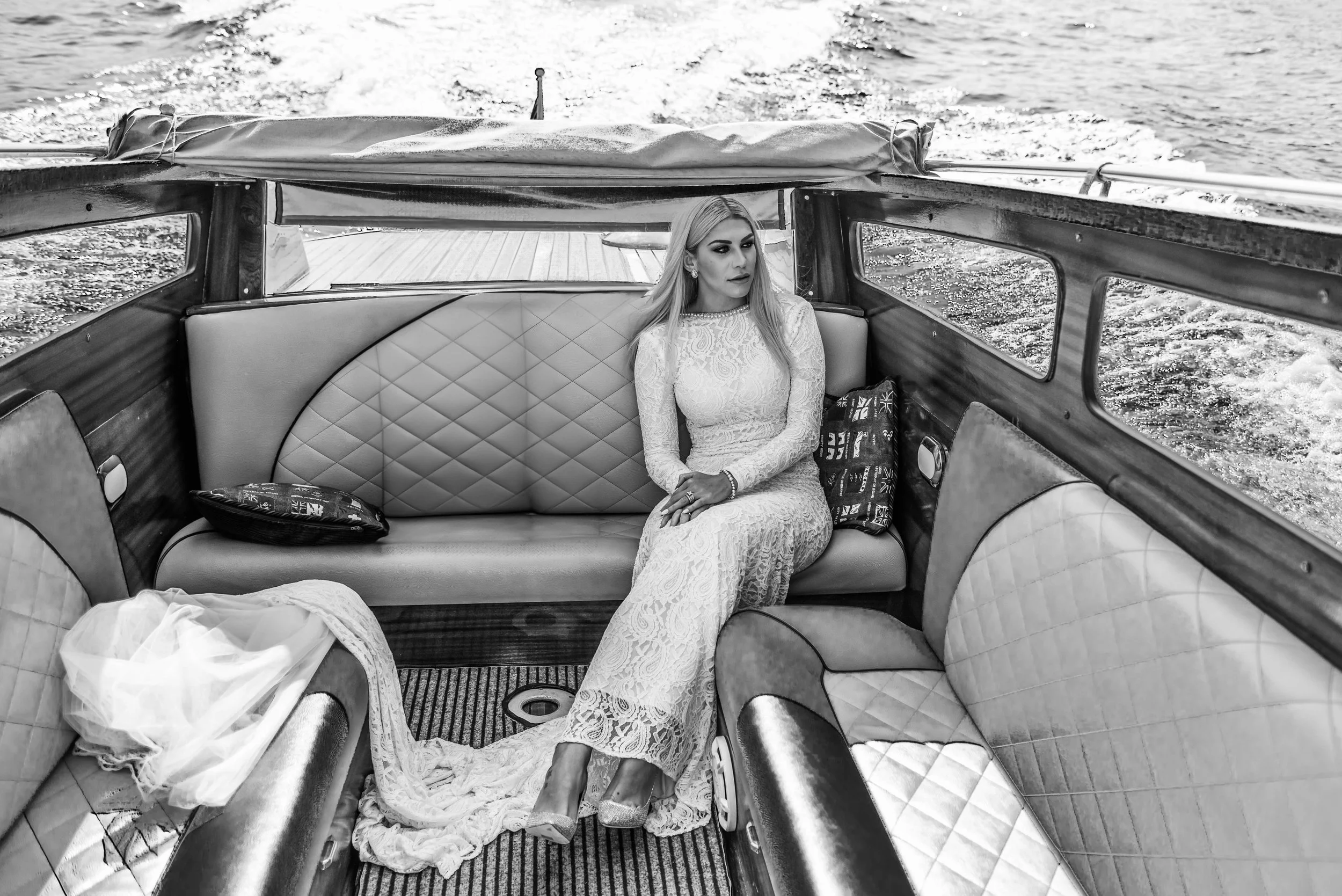 A woman in a long lace dress sitting inside a boat on the water, with patterned pillows around her and looking to the side.
