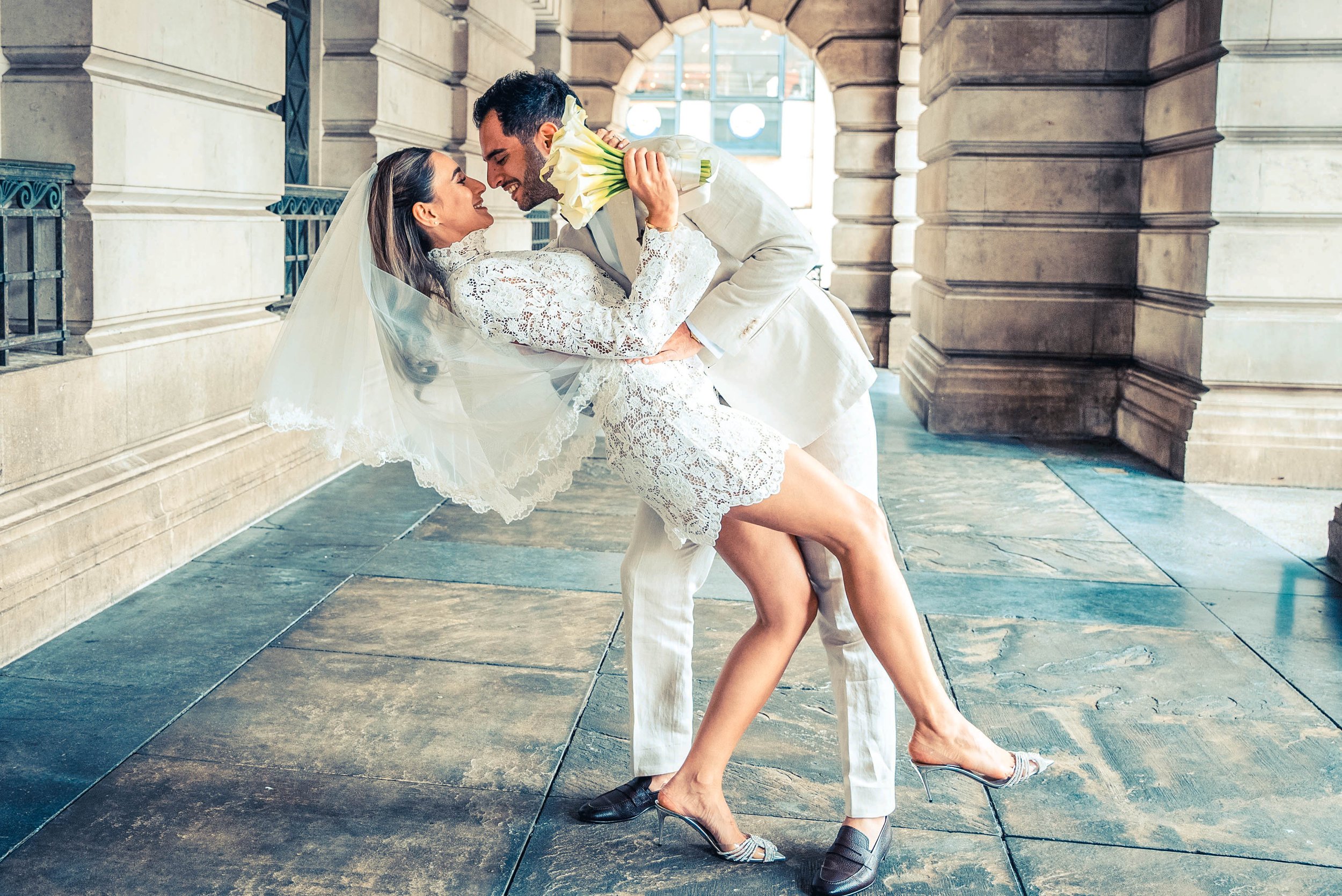 A bride and groom sharing a dance with the groom dipping the bride, who is holding a bouquet, inside a historic building with stone archways and large windows.