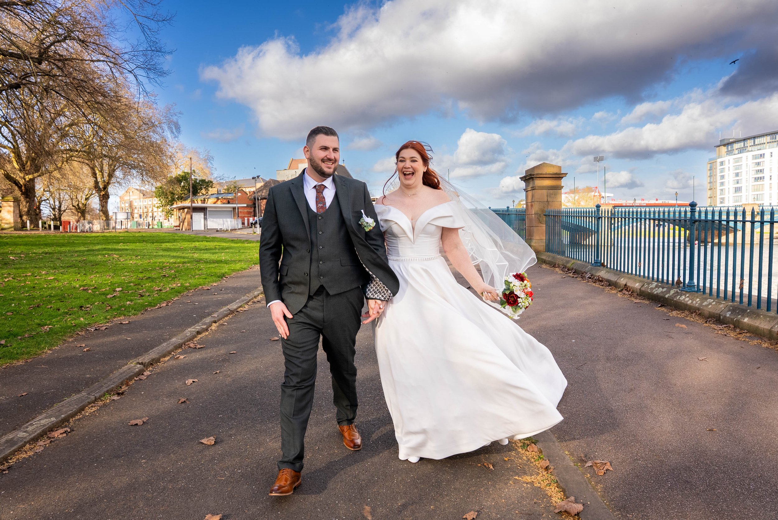 A newlywed couple holding hands and walking outdoors on a paved path, with a lake and city buildings in the background, under a partly cloudy sky.AB Studio Creative ,wedding photographer , Nottingham