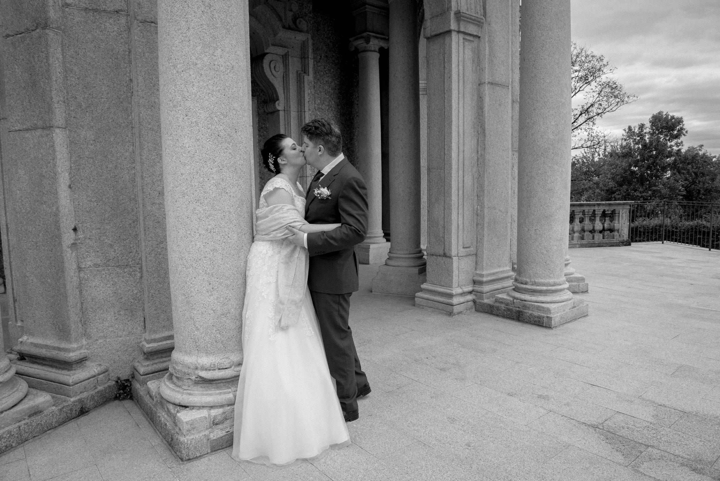 A bride and groom kissing outdoors beside stone columns with trees and a cloudy sky in the background, in black and white.