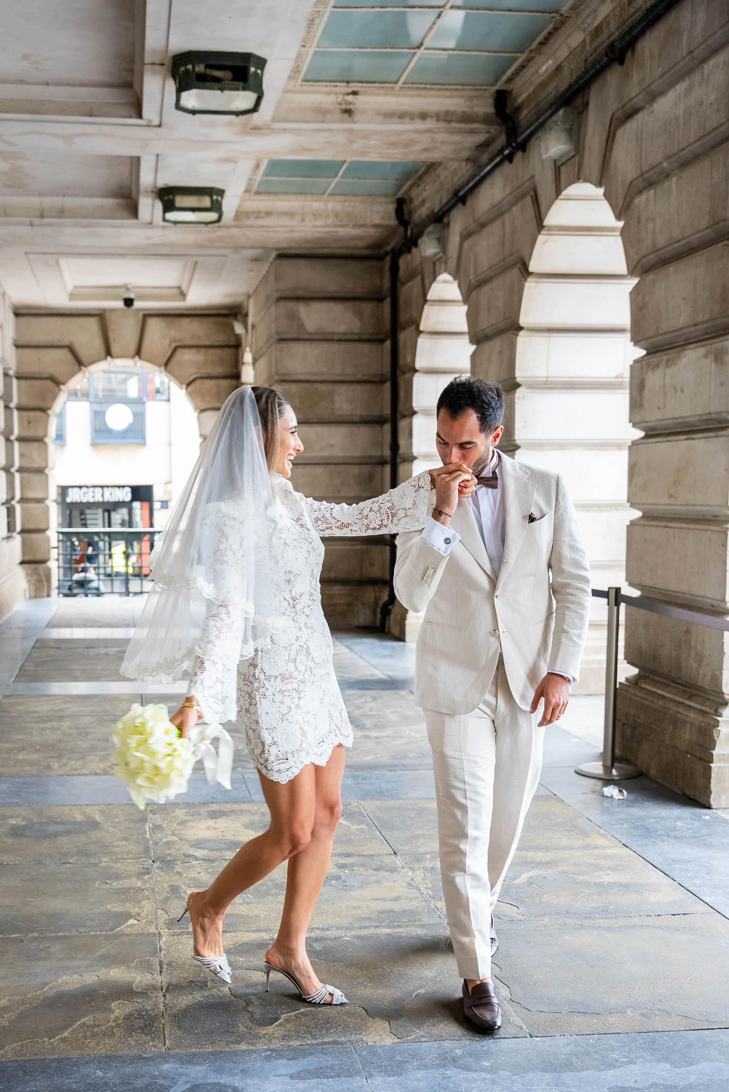 A bride in a lace dress holding a bouquet is kissing a groom in a cream suit on the hand in an outdoor architectural setting.