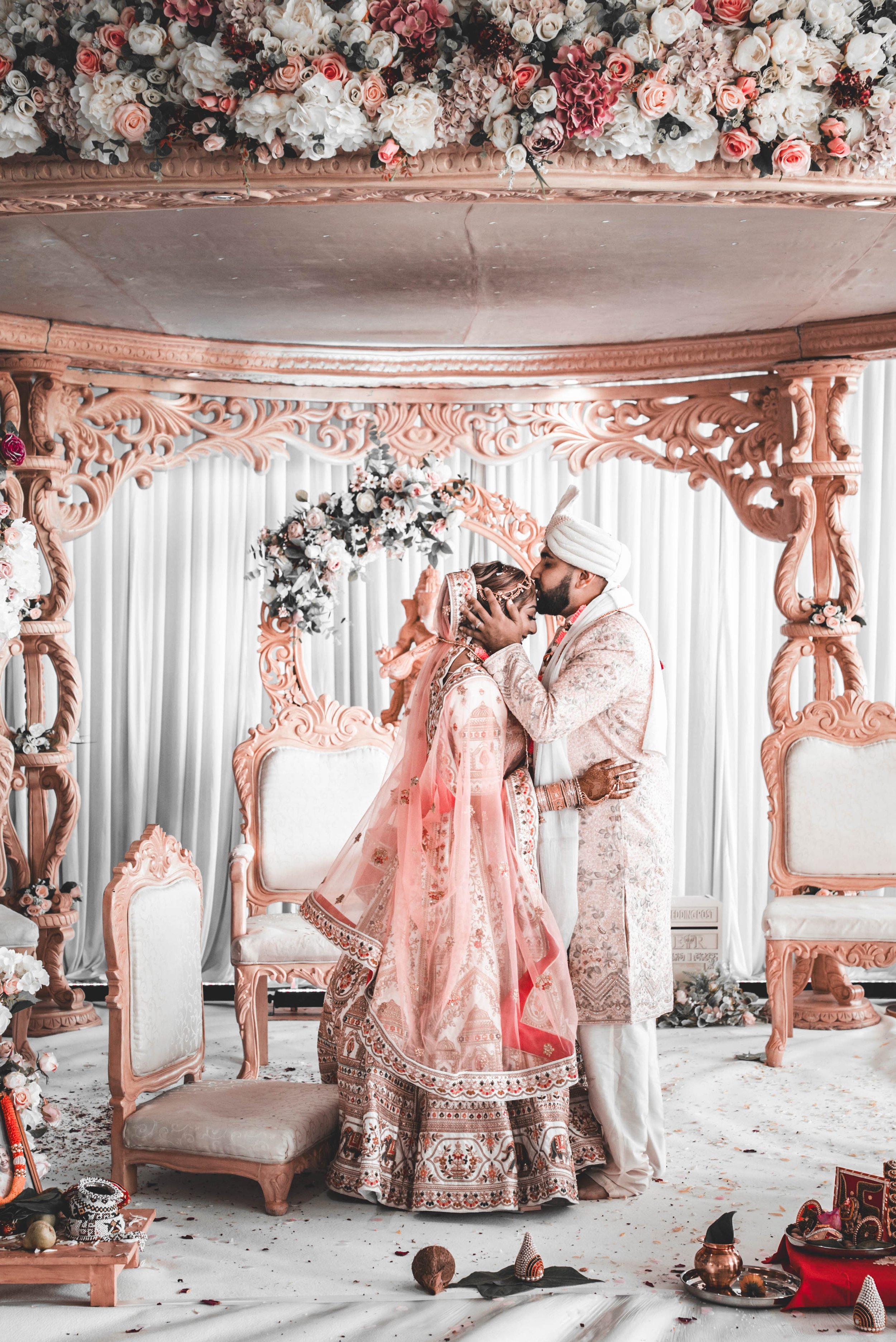 A couple dressed in traditional Indian wedding attire sharing an intimate moment during their wedding ceremony, standing in front of an ornate floral backdrop with pink, white, and red flowers and intricate carved wood details.
