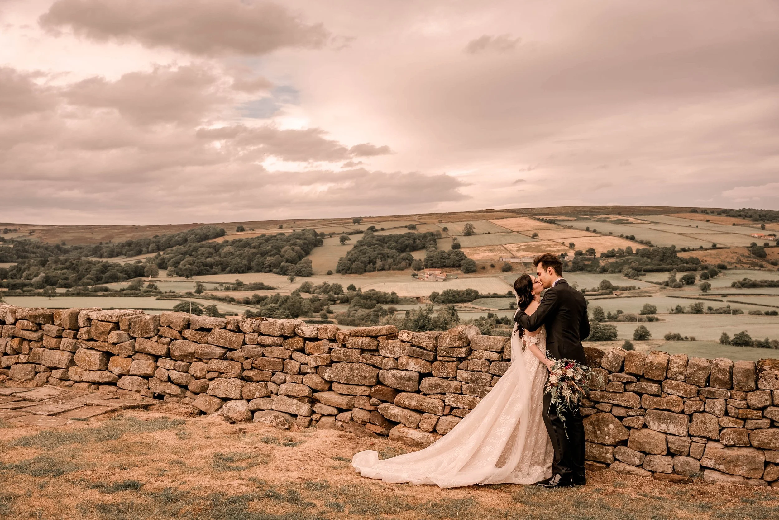 A bride and groom standing close together behind a stone wall, sharing a kiss against a rural landscape with rolling hills and cloudy sky.