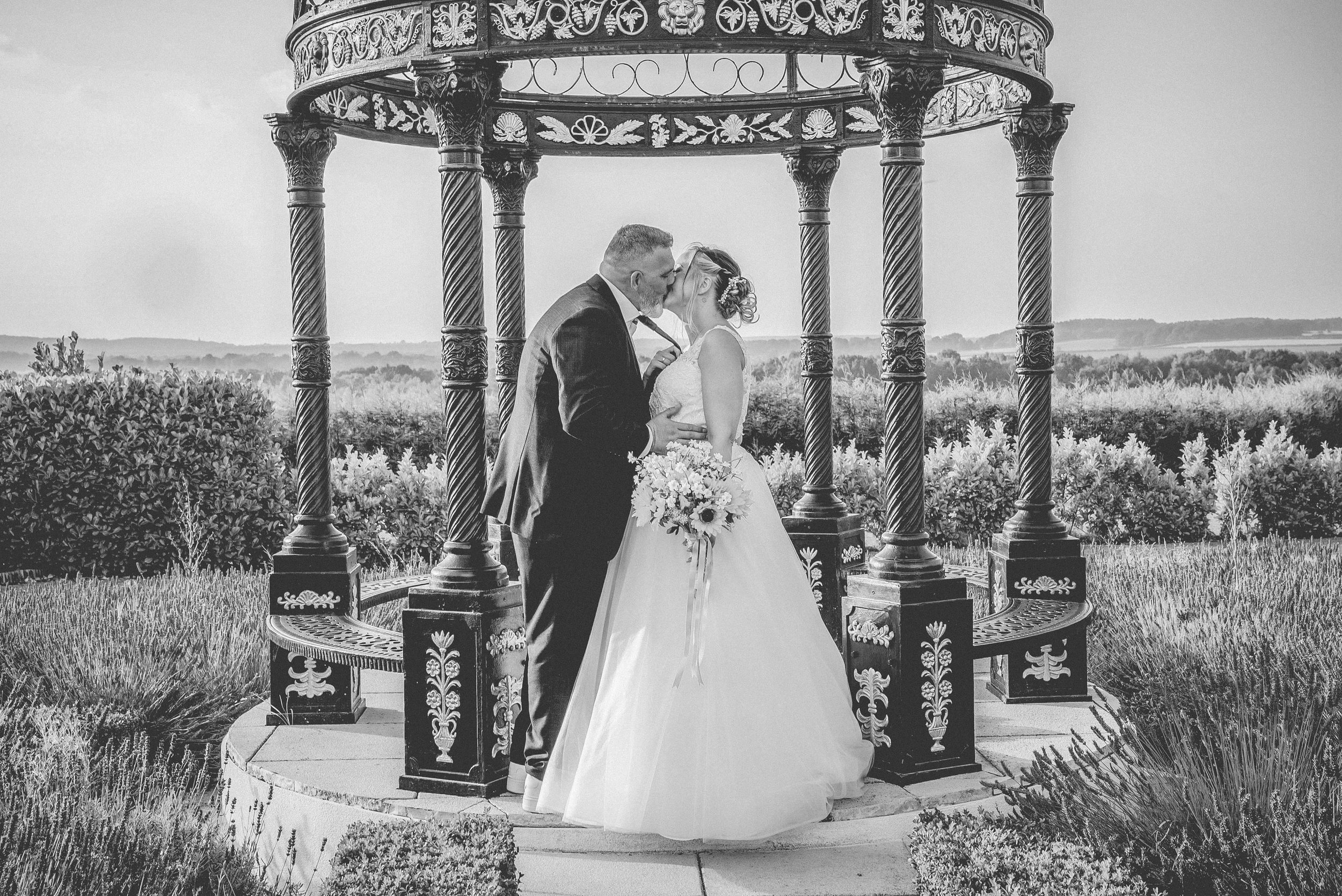 Black and white photo of a bride and groom kissing under an ornate gazebo outdoors, with a scenic landscape in the background.