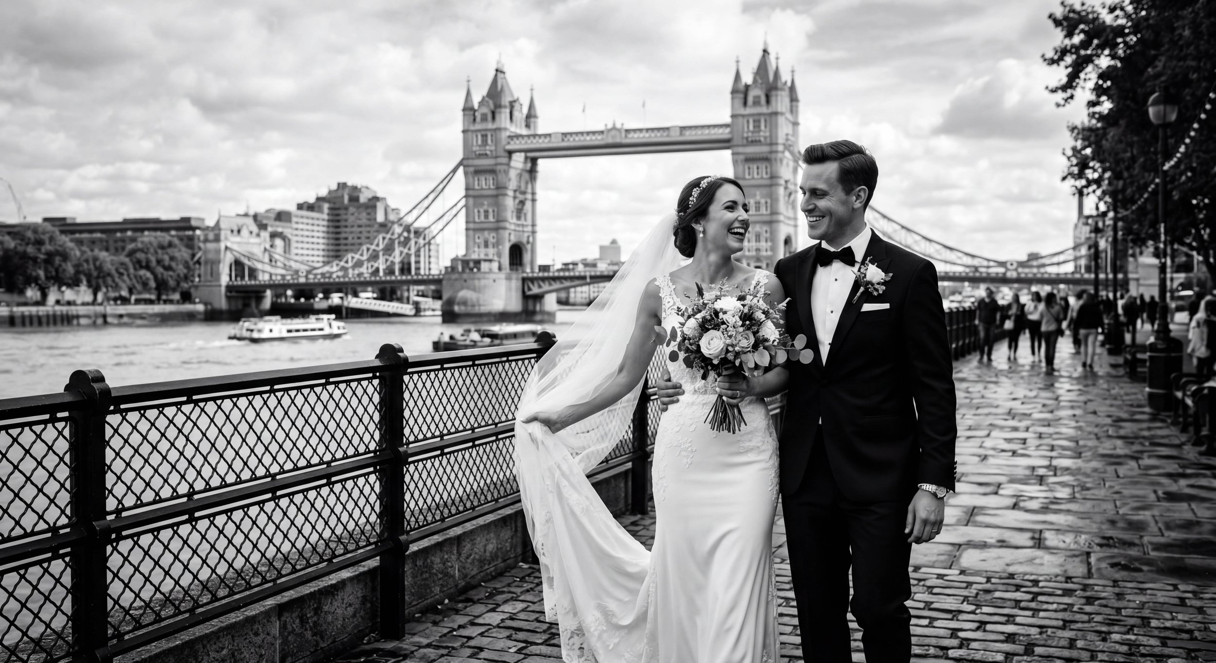 Black and white photo of a bride and groom smiling and walking together near the Tower Bridge in London. The bride is holding a bouquet and wearing a wedding dress with a veil, while the groom is dressed in a tuxedo.