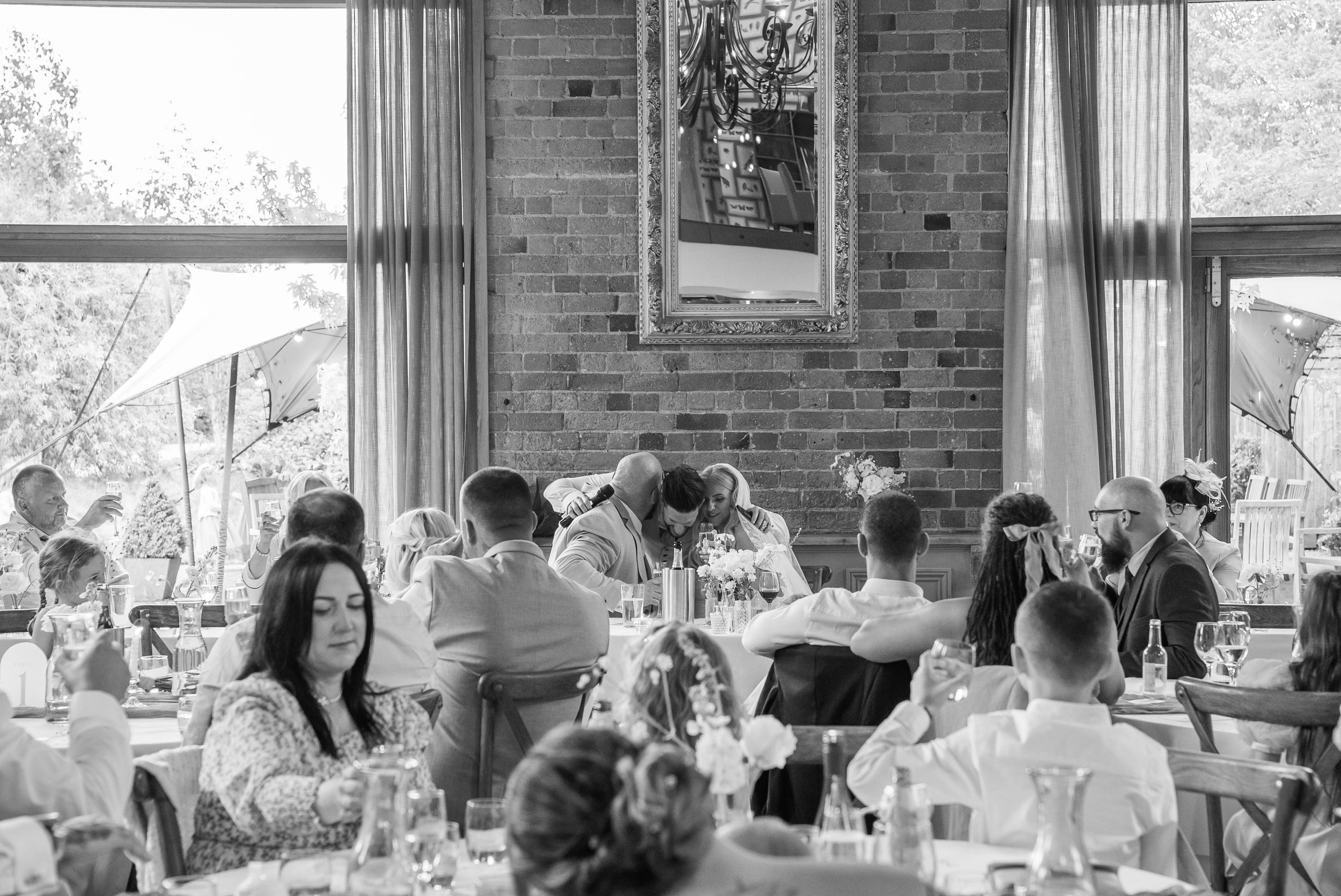 Guests at a wedding reception seated at tables, with a brick wall, curtains, and windows in the background.