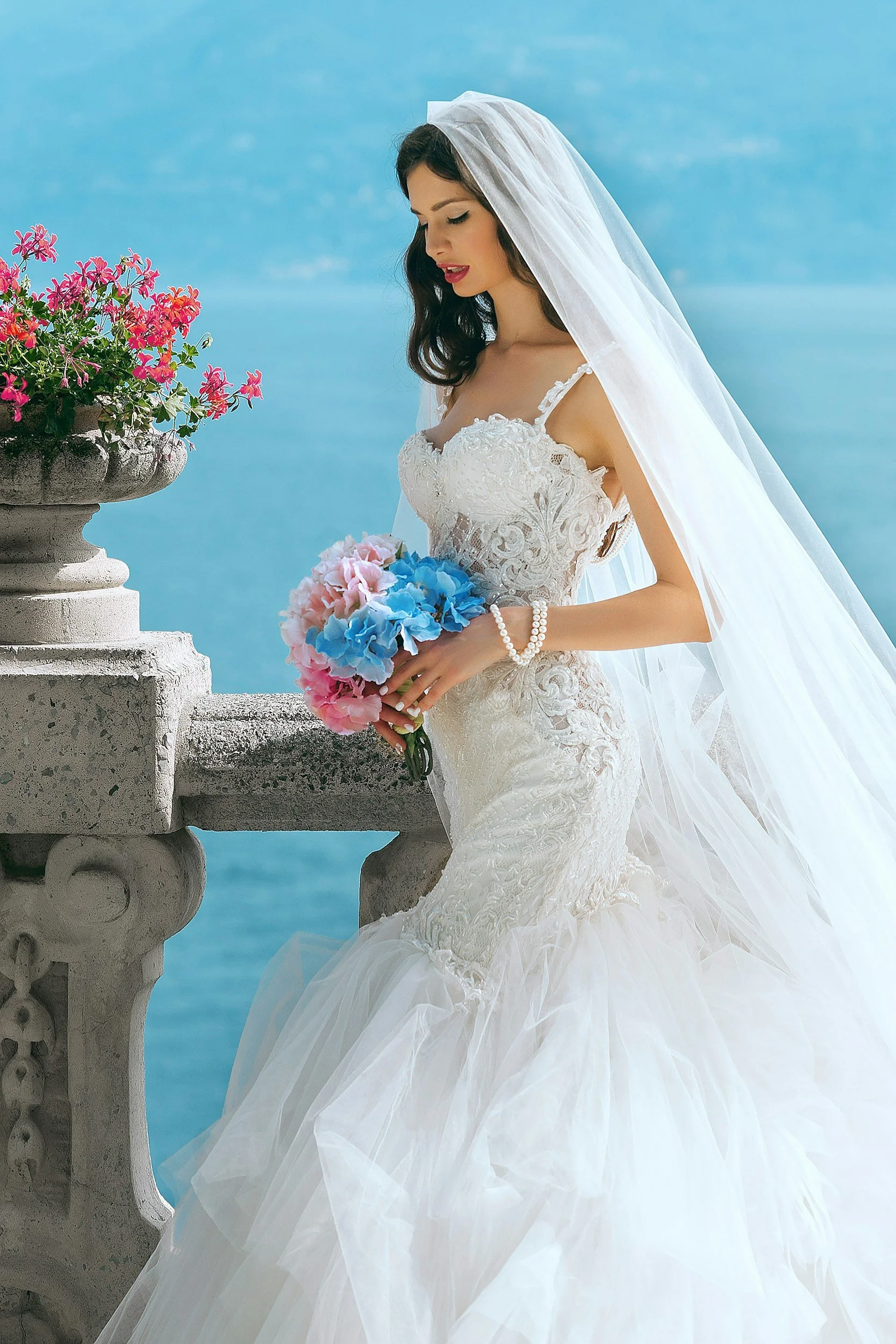 A bride in a white wedding dress with lace details and a long veil stands on a balcony holding a bouquet of pink, blue, and white flowers, with a large planter of pink flowers nearby and a sea or lake in the background.