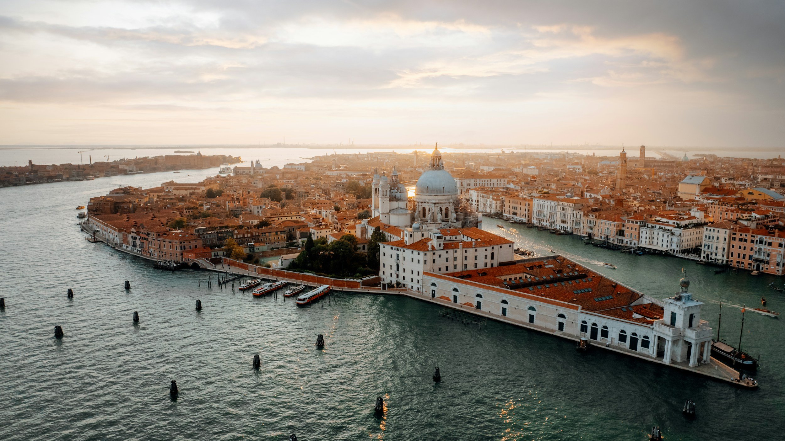 Aerial view of Venice, Italy, showing historic buildings, waterways, boats, and the dome of a church during sunset.