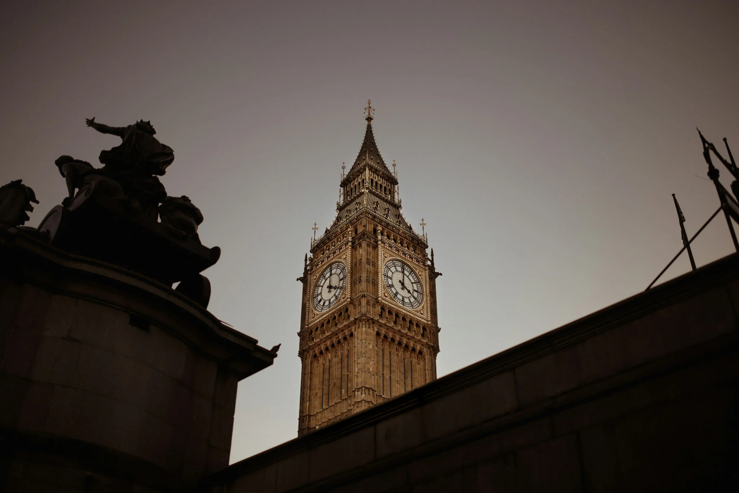 A low-angle view of Big Ben clock tower in London, with a dark silhouette of a statue in the foreground and a pale sky in the background.