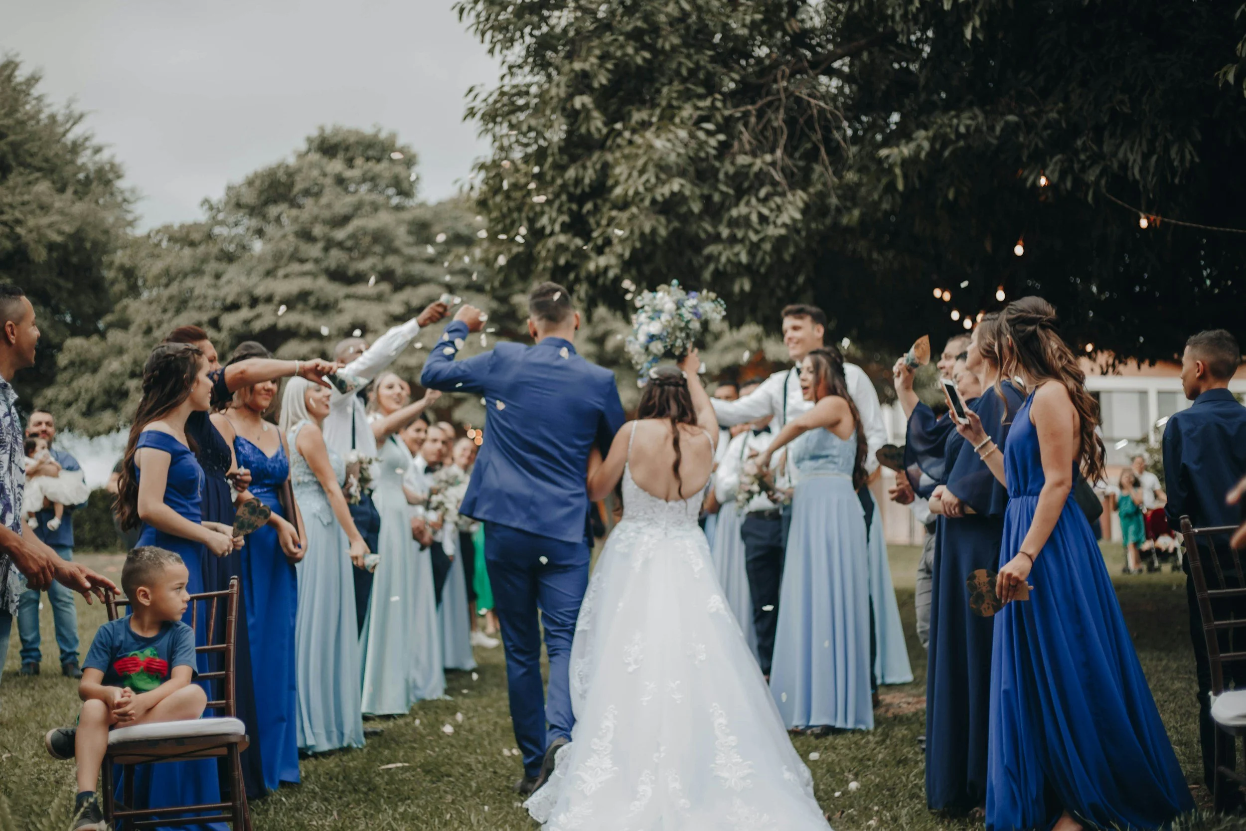 A bride and groom dancing outdoors at a wedding reception, surrounded by guests in blue and light blue dresses and suits, with some throwing flower petals, under a large tree with string lights.
