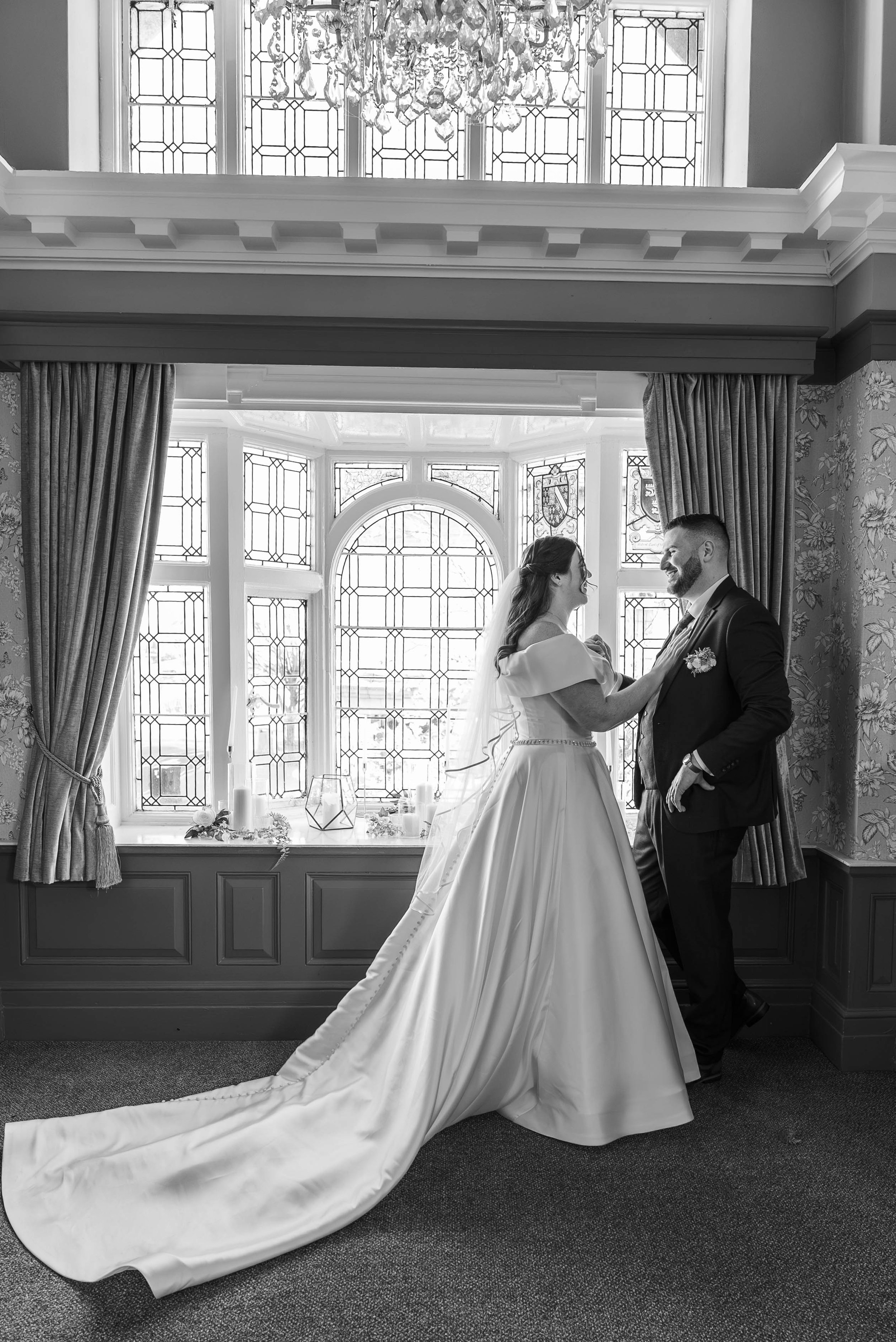 A bride and groom dancing in a room with large stained glass windows and ornate curtains. The bride is wearing a strapless wedding gown with a long train, and the groom is in a dark suit. They are smiling at each other.