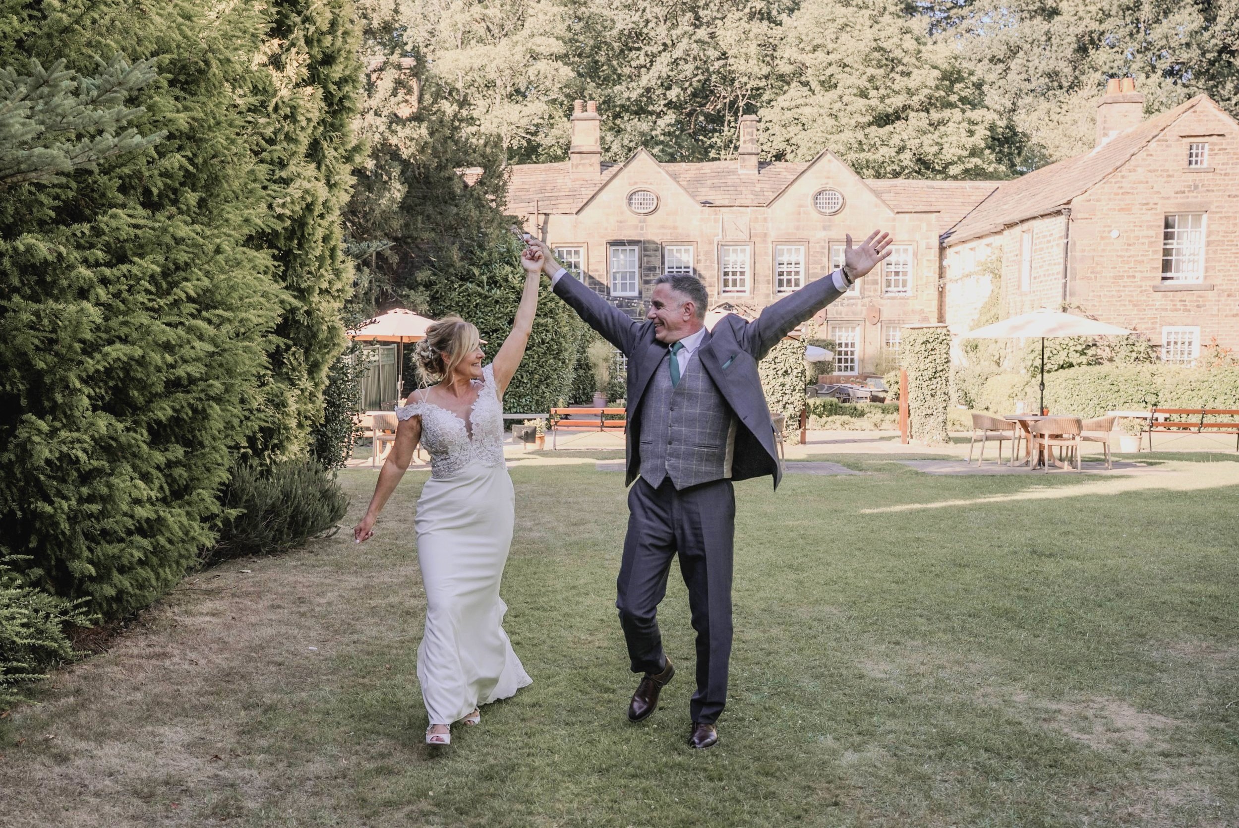 A bride in a white wedding dress and a groom in a suit dancing outdoors in front of a large house, with trees and tables in the background.