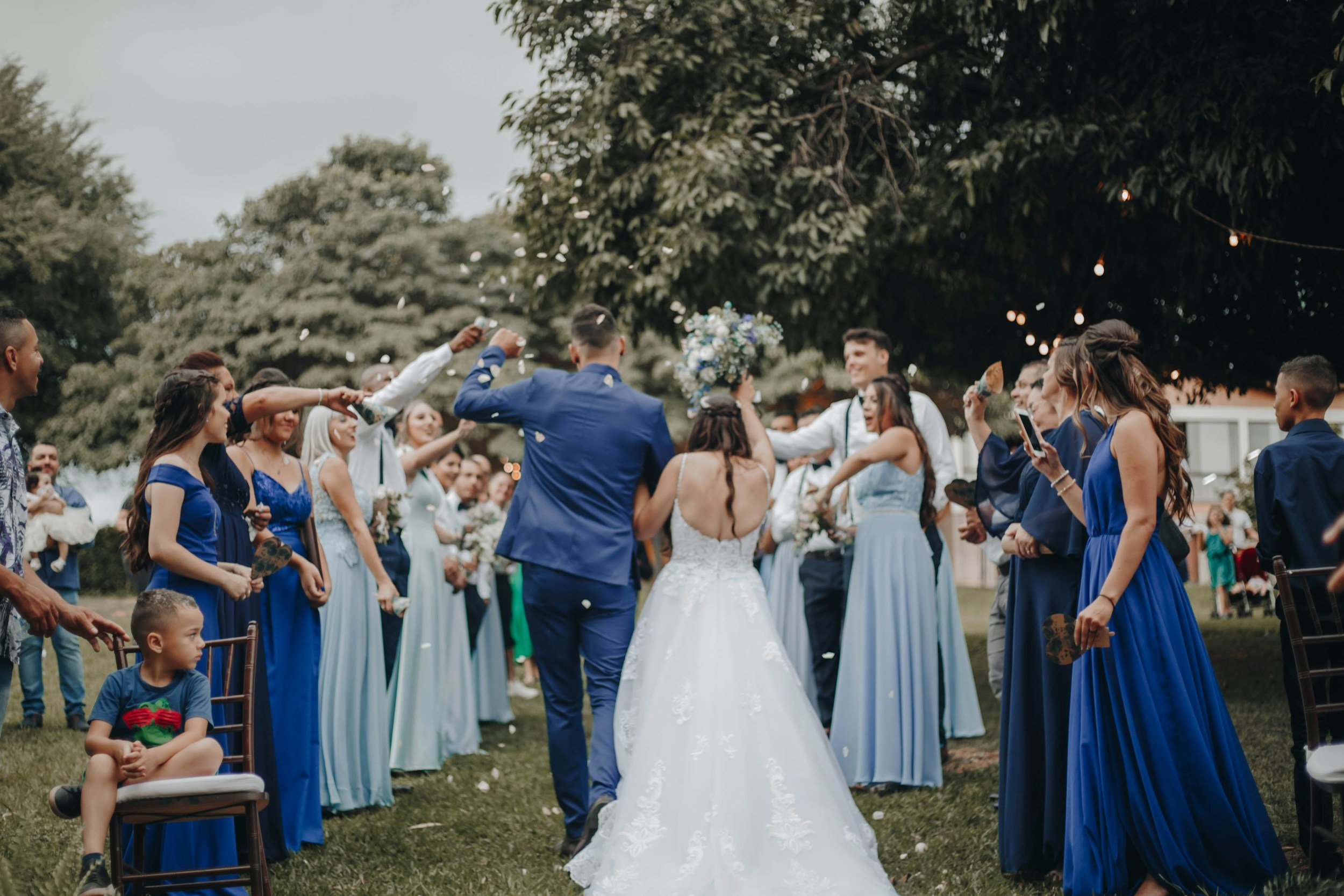 A bride and groom walking together under a shower of confetti, surrounded by friends and family at an outdoor wedding celebration in a lush garden setting.