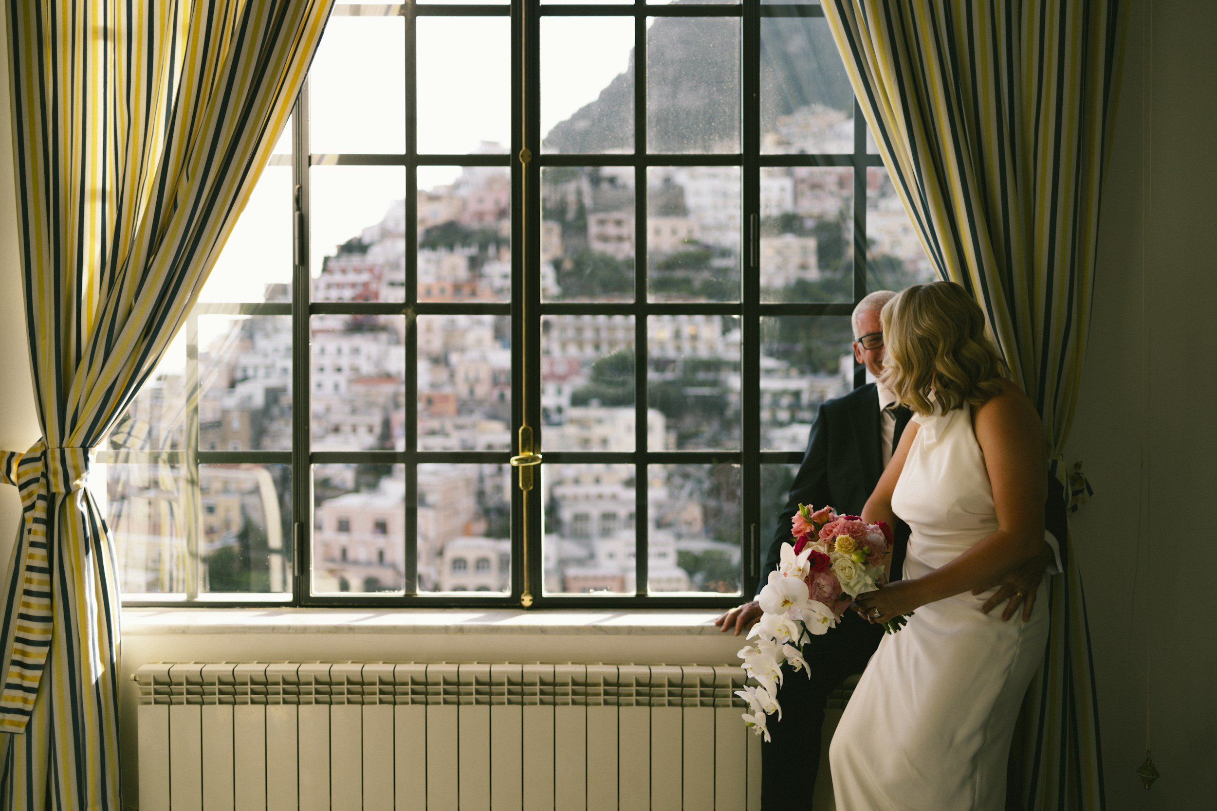 A bride in a white wedding dress holding a bouquet of pink and white flowers, standing near a window with striped curtains, with a man in a suit behind her, and a cityscape visible outside.