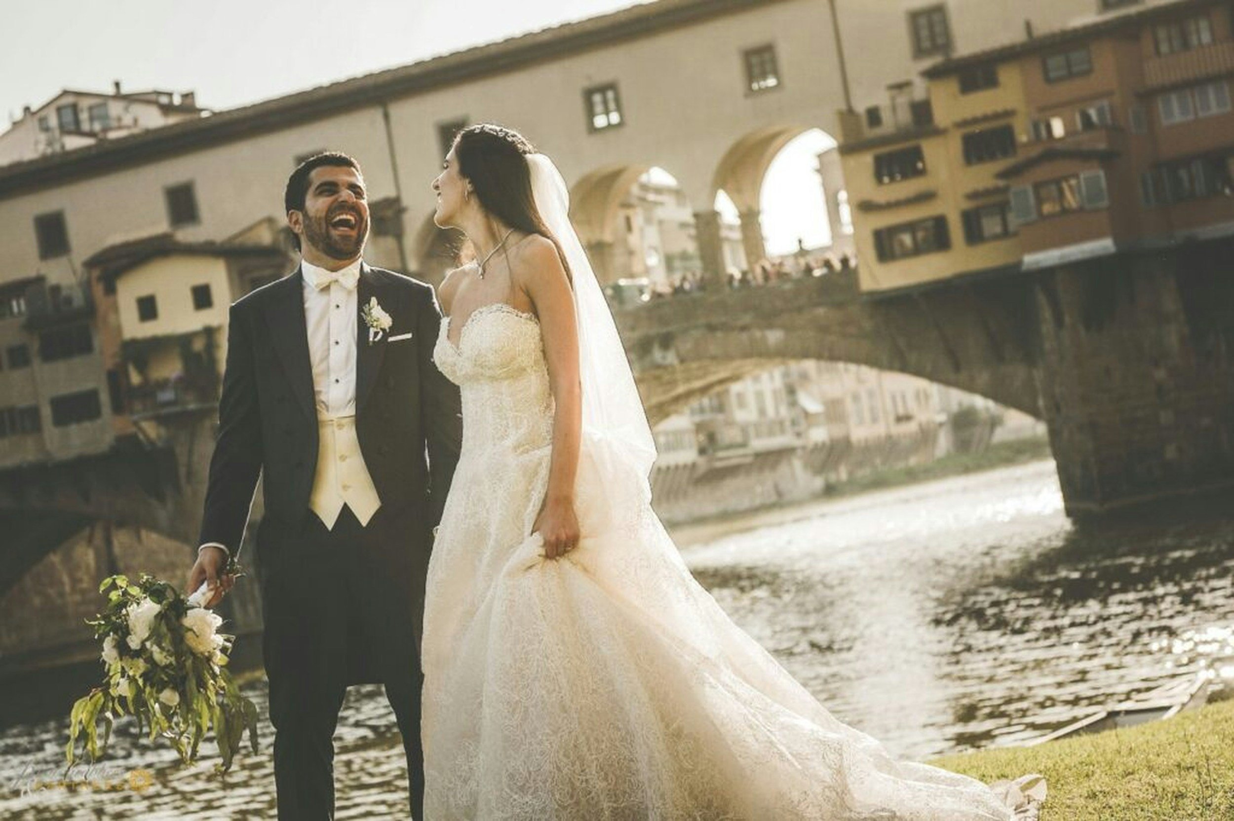 A newlywed couple in wedding attire enjoying a moment by the river with an old stone bridge and colorful buildings in the background.