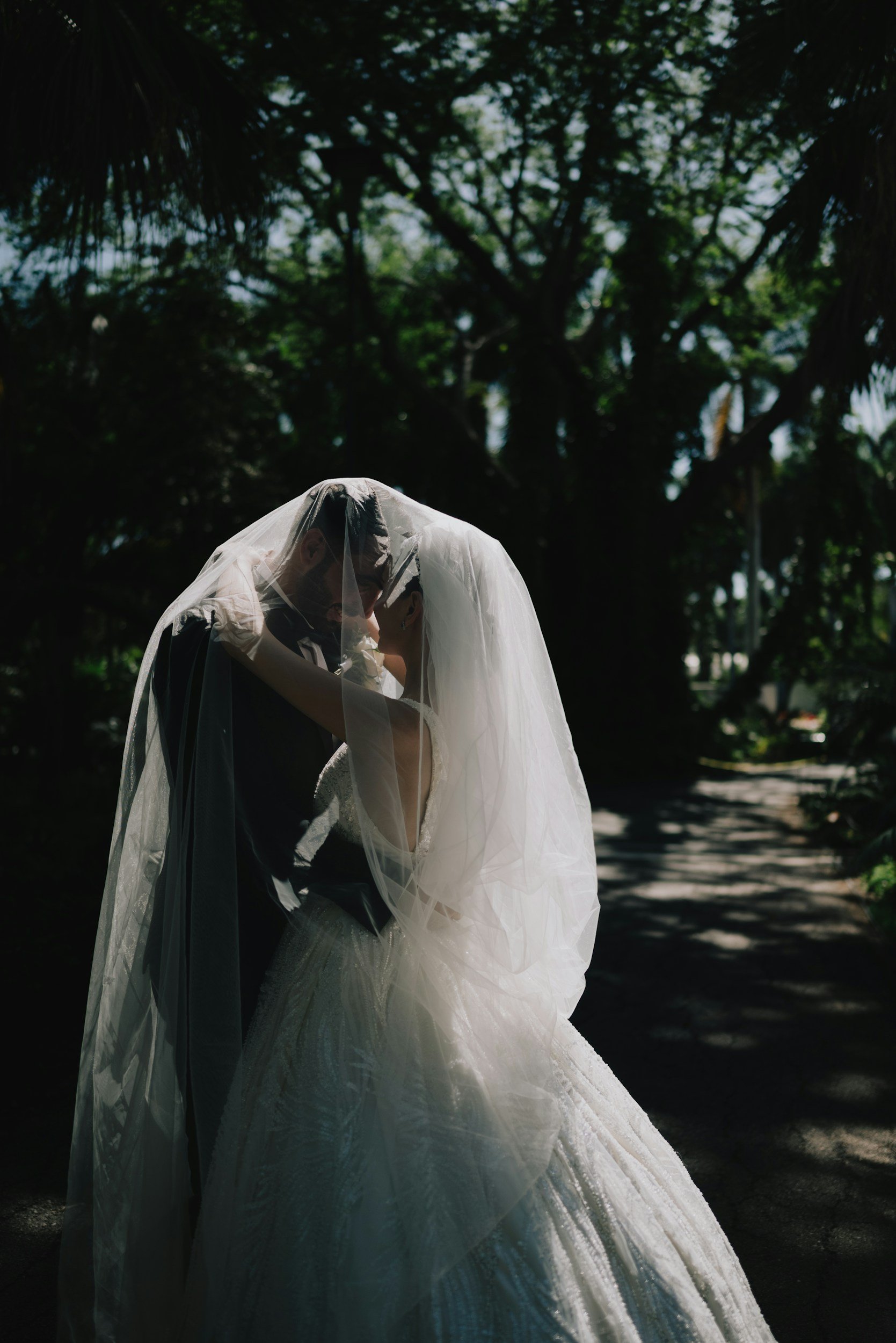 A bride and groom embrace outdoors, with the bride wearing a white wedding gown and veil, and the groom dressed formally, surrounded by trees and natural light.