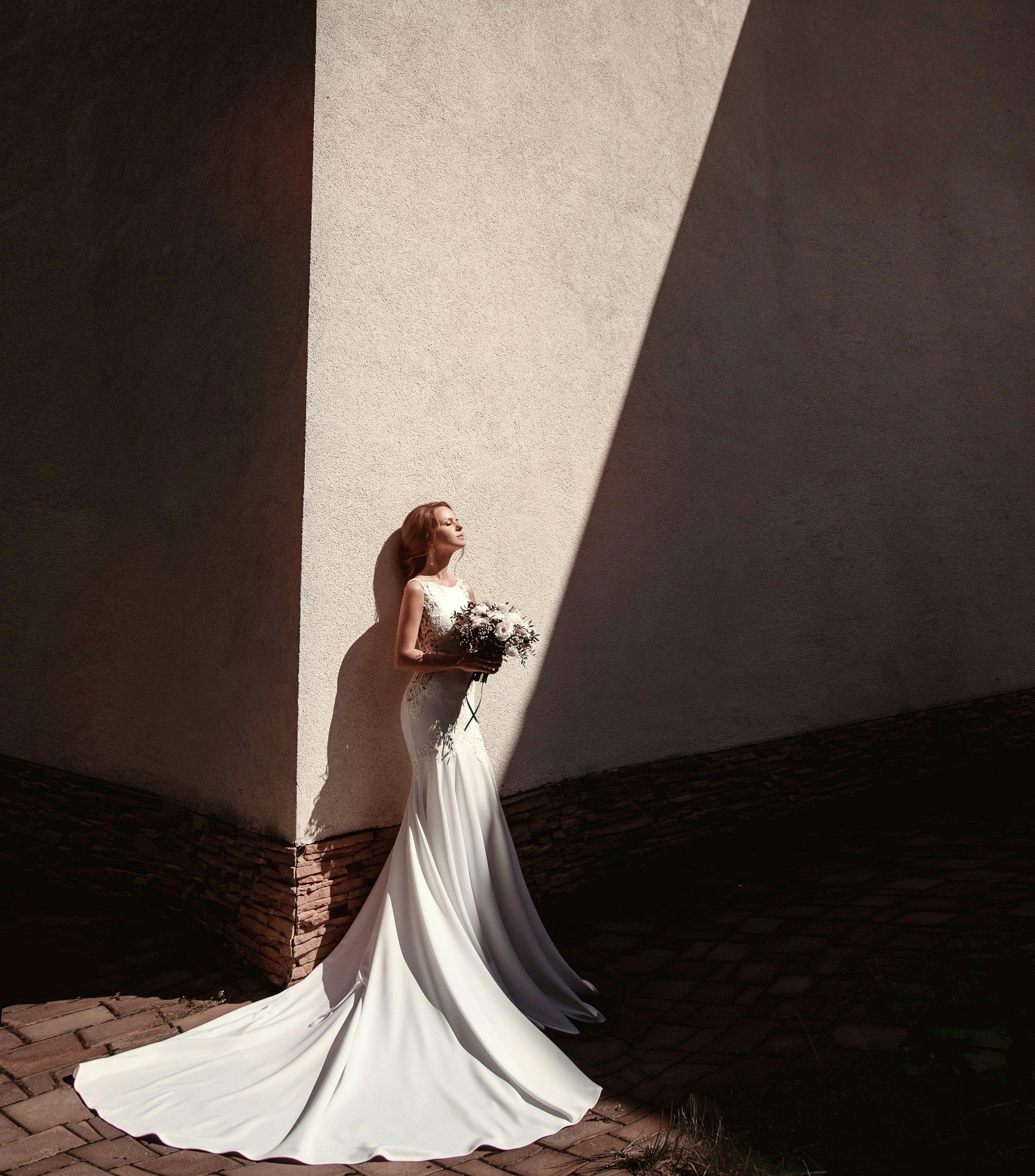 A woman in a white wedding dress with a long train, holding a bouquet of flowers, standing against a wall with dramatic shadows cast by sunlight.